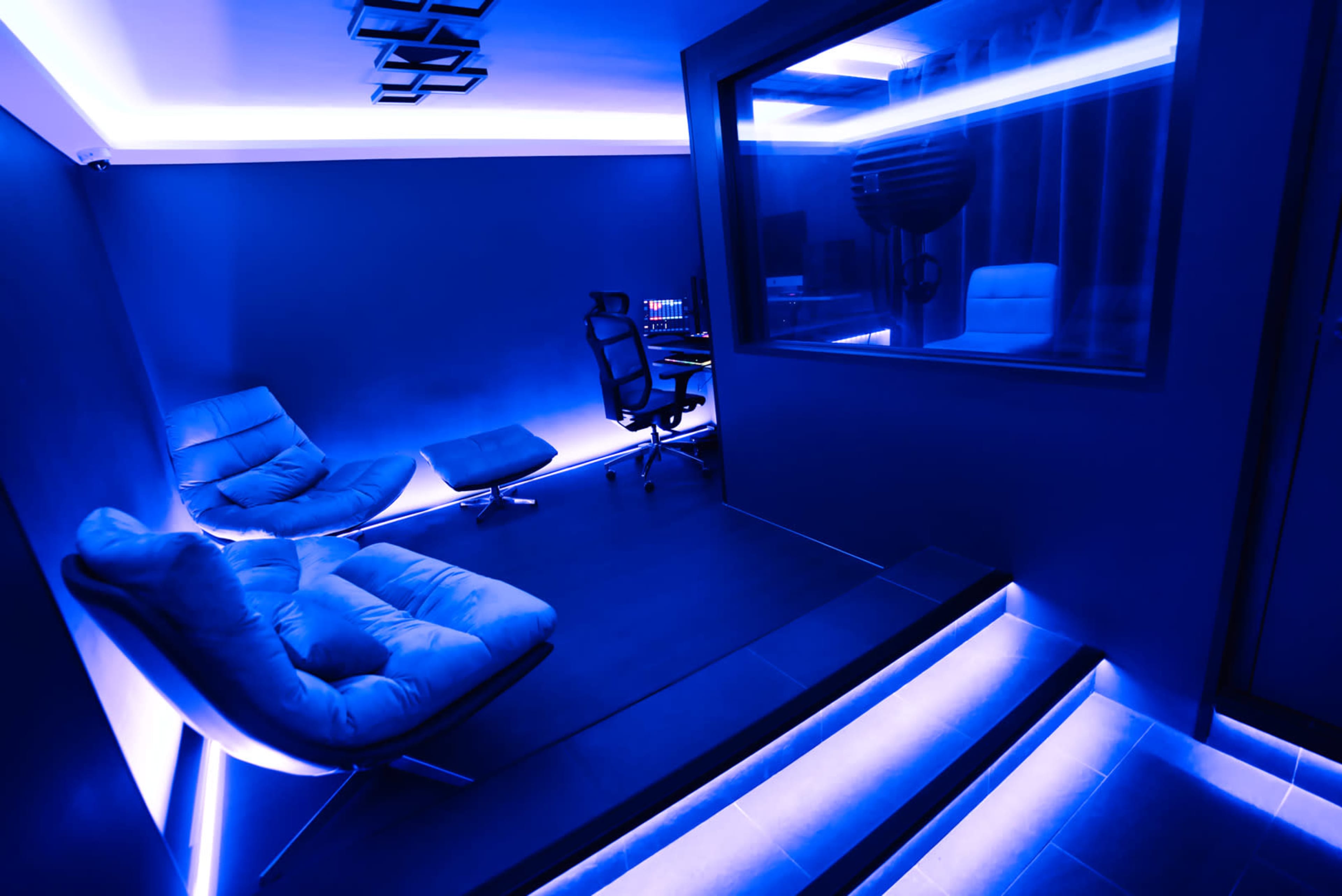 A minimalist recording studio featuring blue lighting, a comfortable seating area, and a separate soundproof room.