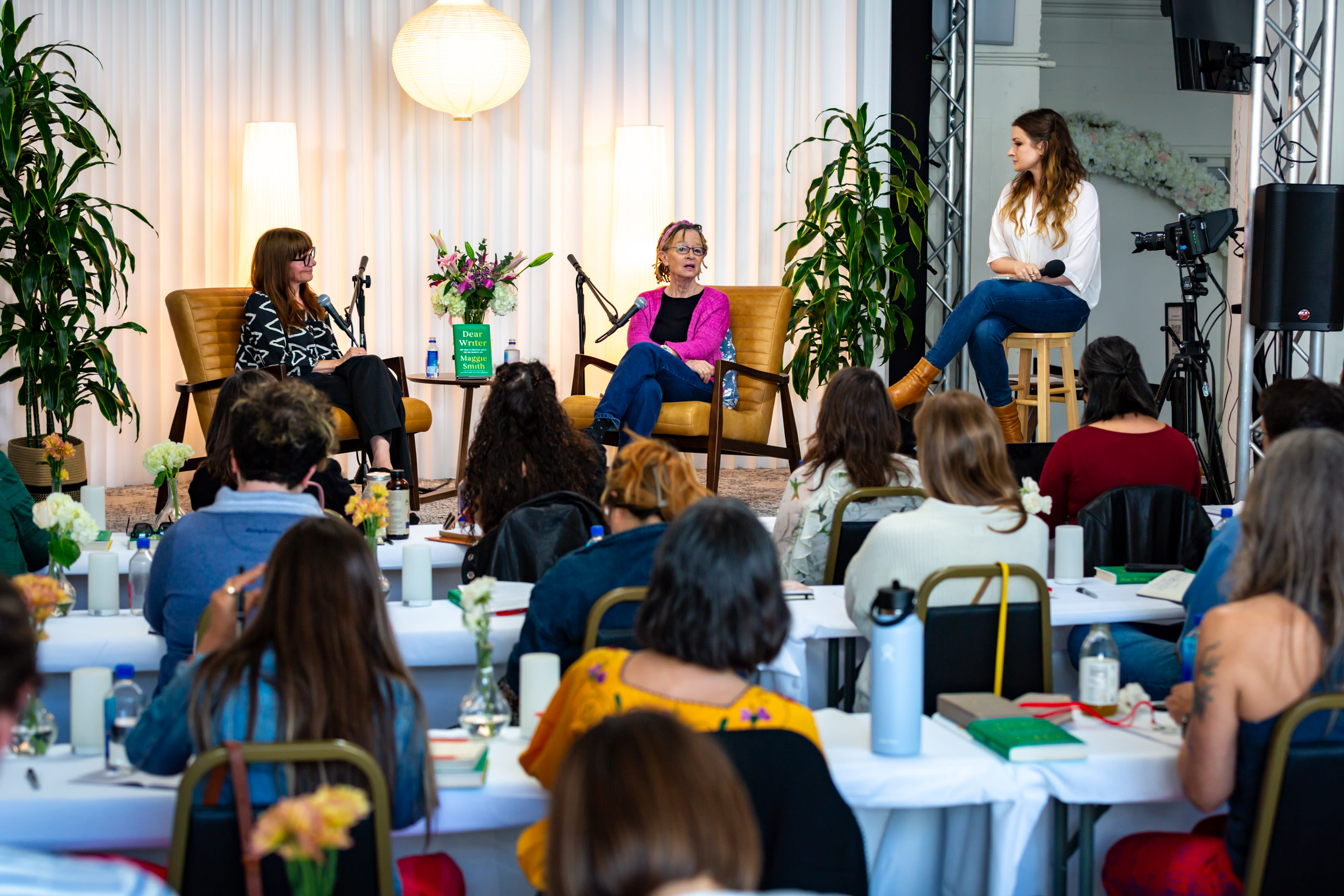 A panel discussion is taking place with three women seated on stage, while an audience watches attentively from their tables filled with notebooks and water bottles.