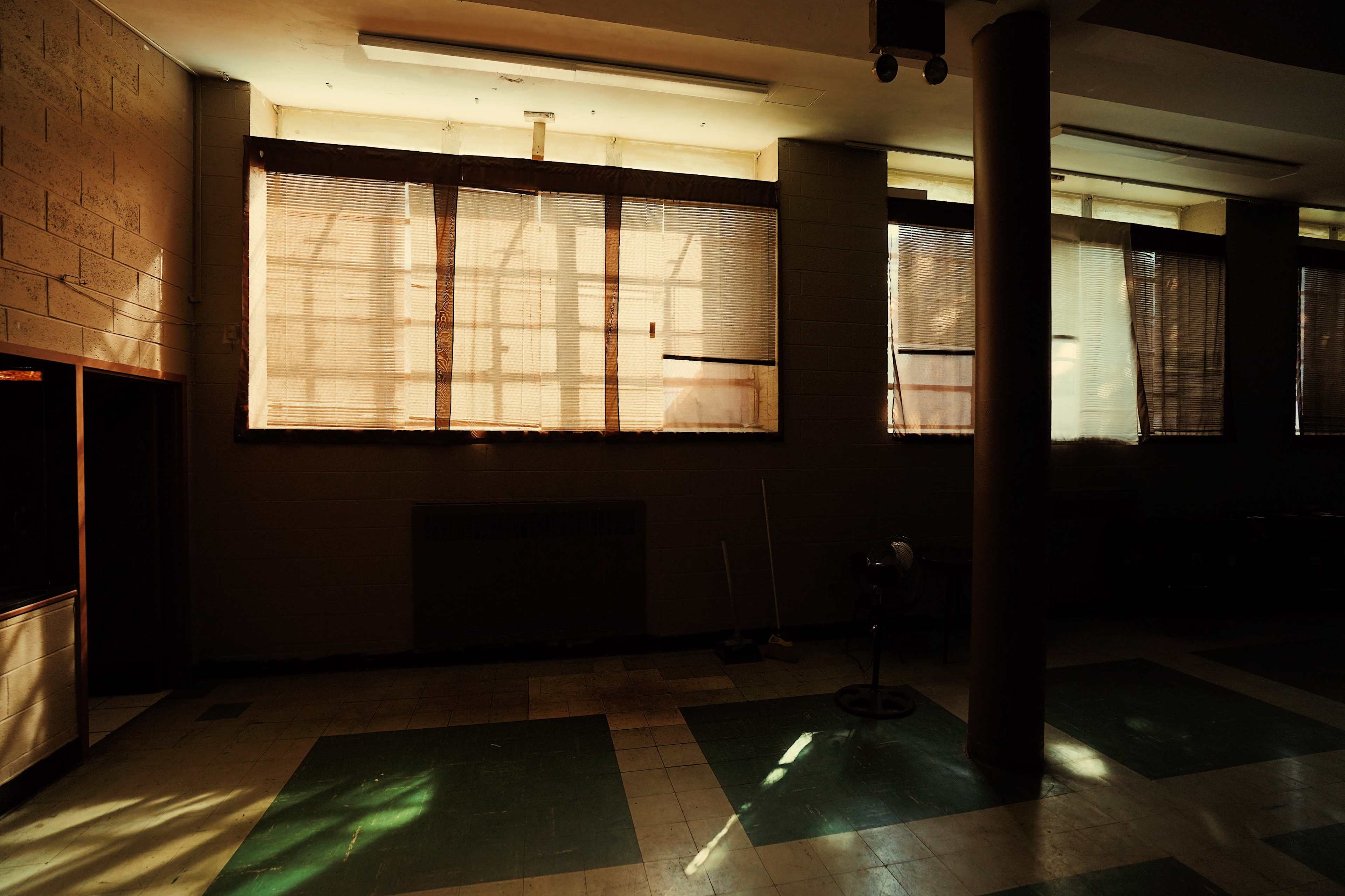 A dimly lit interior space with large windows partially covered by blinds, casting shadows on the green and white tiled floor.