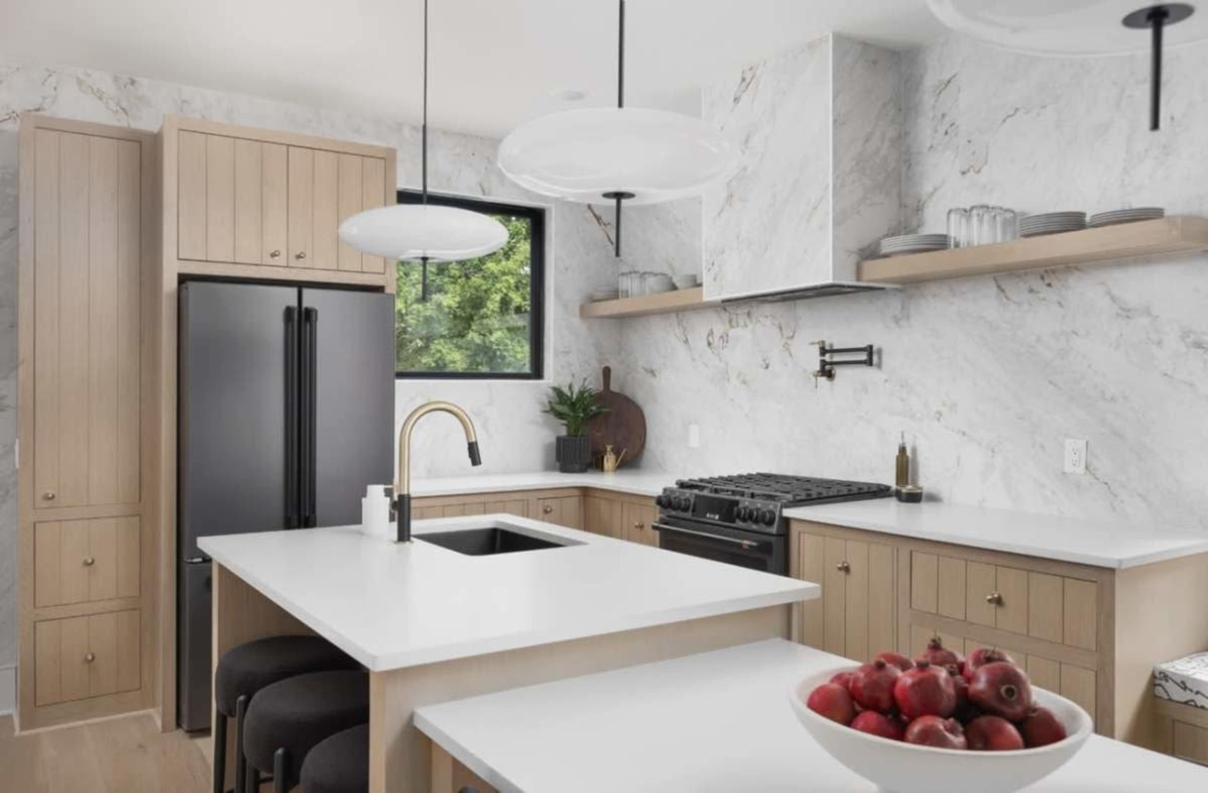 The image shows a modern kitchen featuring a black refrigerator, a gas range, a marble backsplash, wooden cabinetry, and a central island with a fruit bowl.