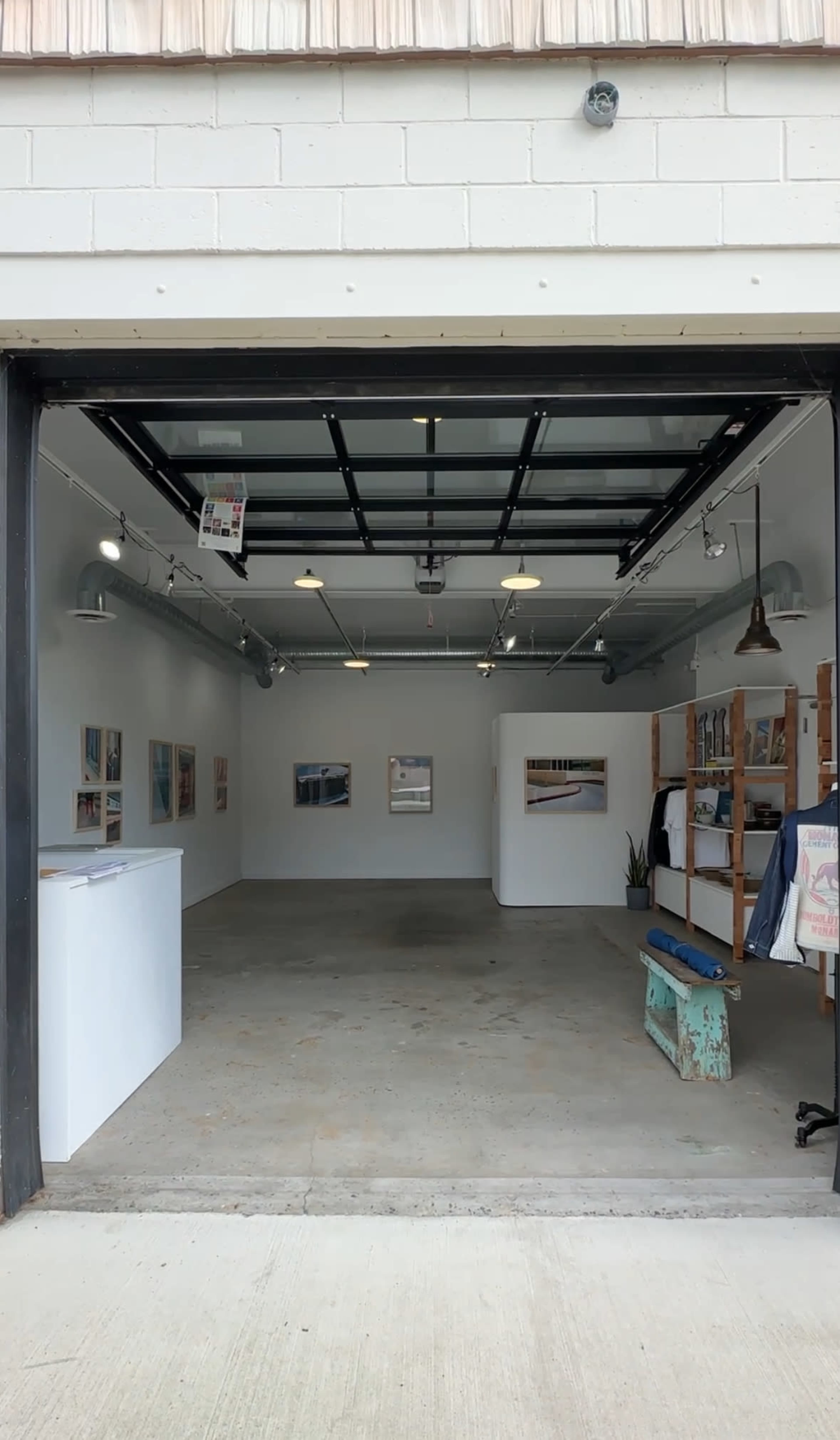 The image shows an open art gallery space with white walls, artwork displayed on the walls, and furnishings such as a bench and shelving units.