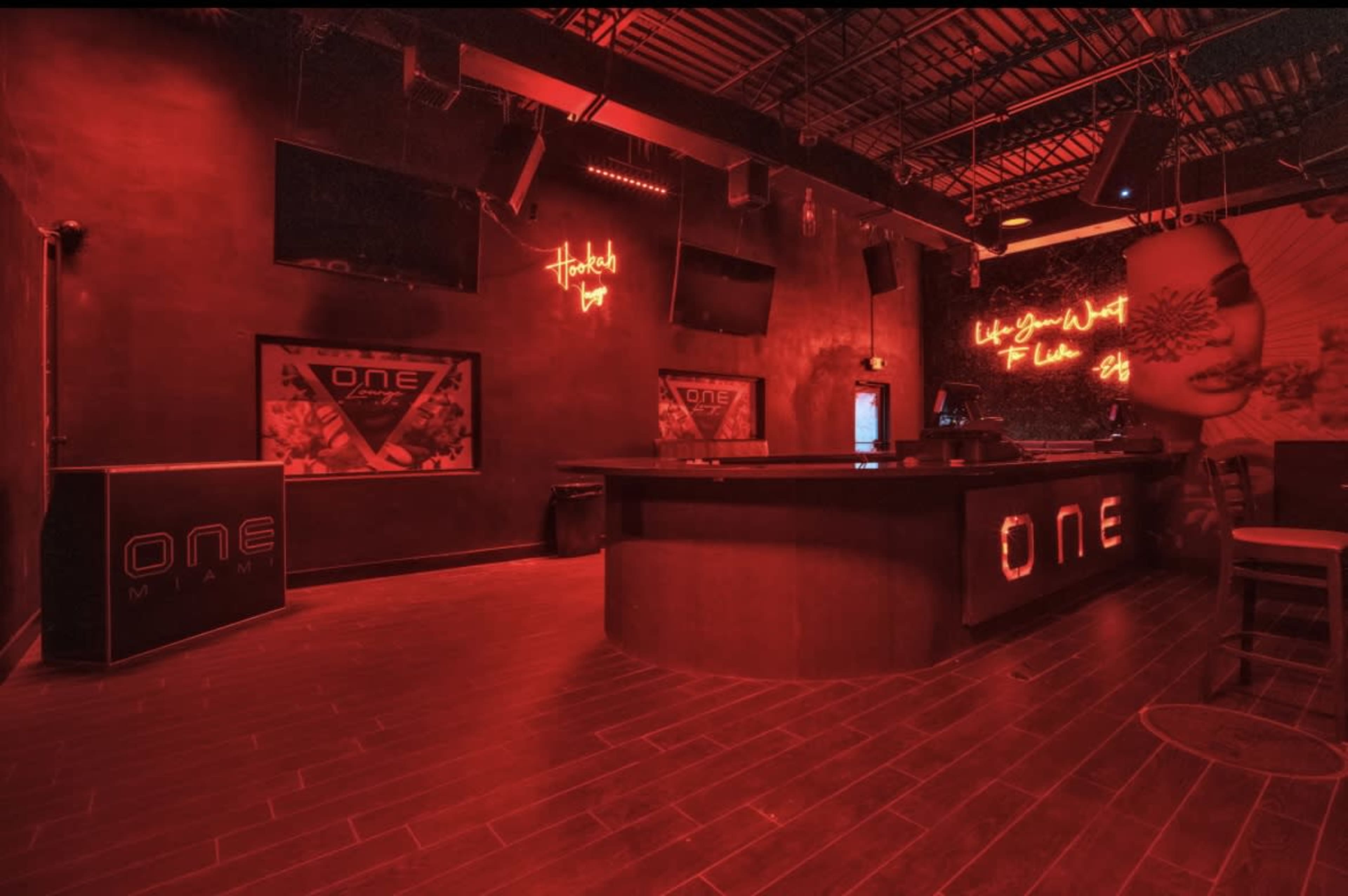 The image shows the interior of a dimly lit bar with red lighting, featuring a prominent "ONE" logo on the bar counter and several screens on the walls.