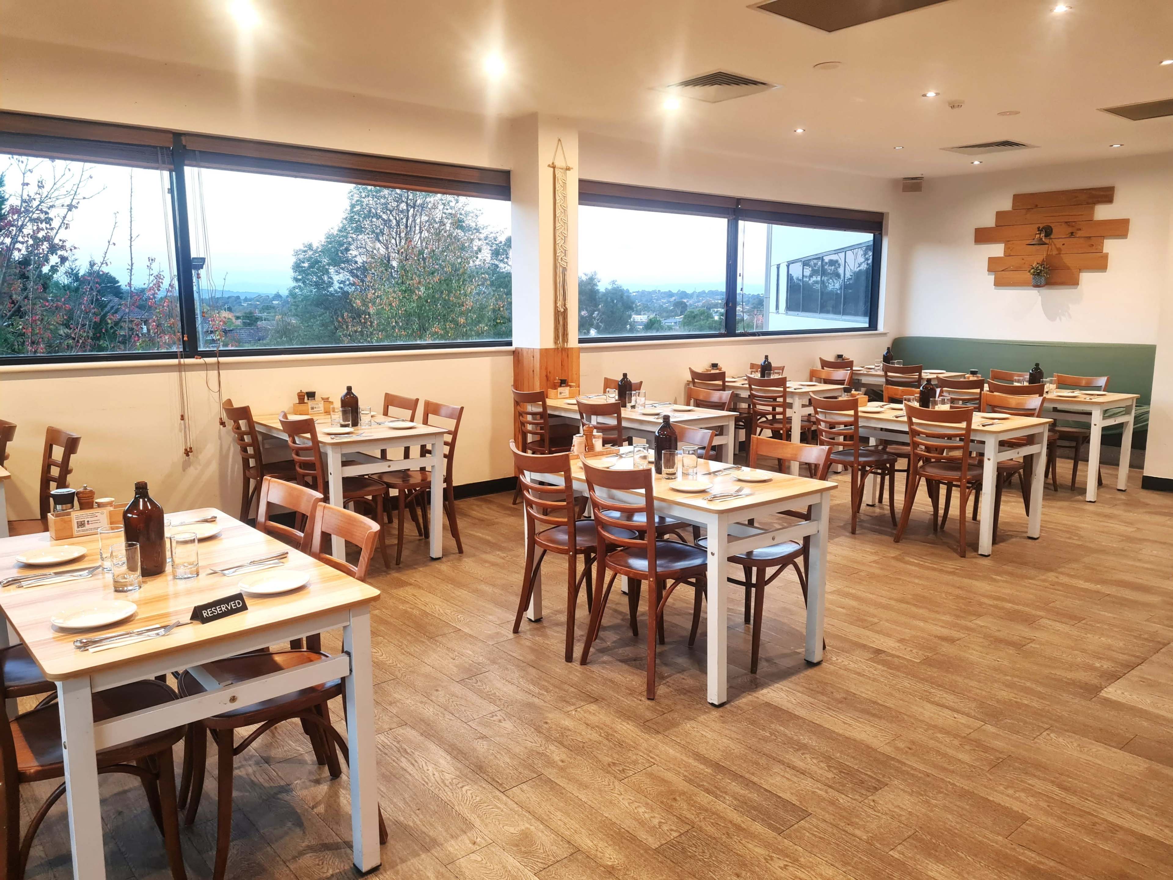 The image shows a restaurant interior with several tables and chairs arranged neatly, large windows offering a view of the outdoors, and a wooden decor on the wall.