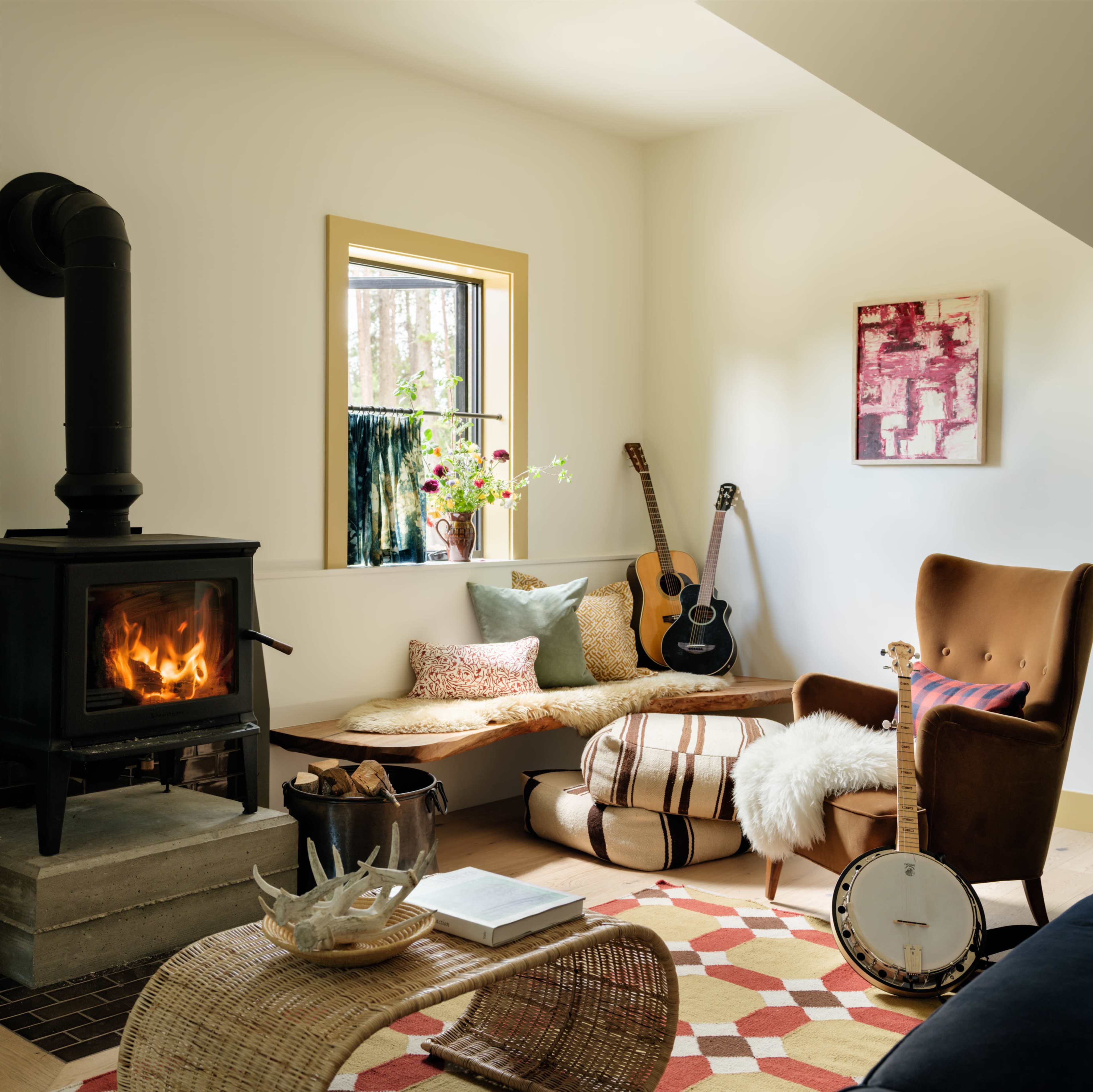 The image shows a cozy living space with a wood-burning stove, a window featuring a vase of flowers, and musical instruments placed around a seating area with decorative pillows and a patterned rug.