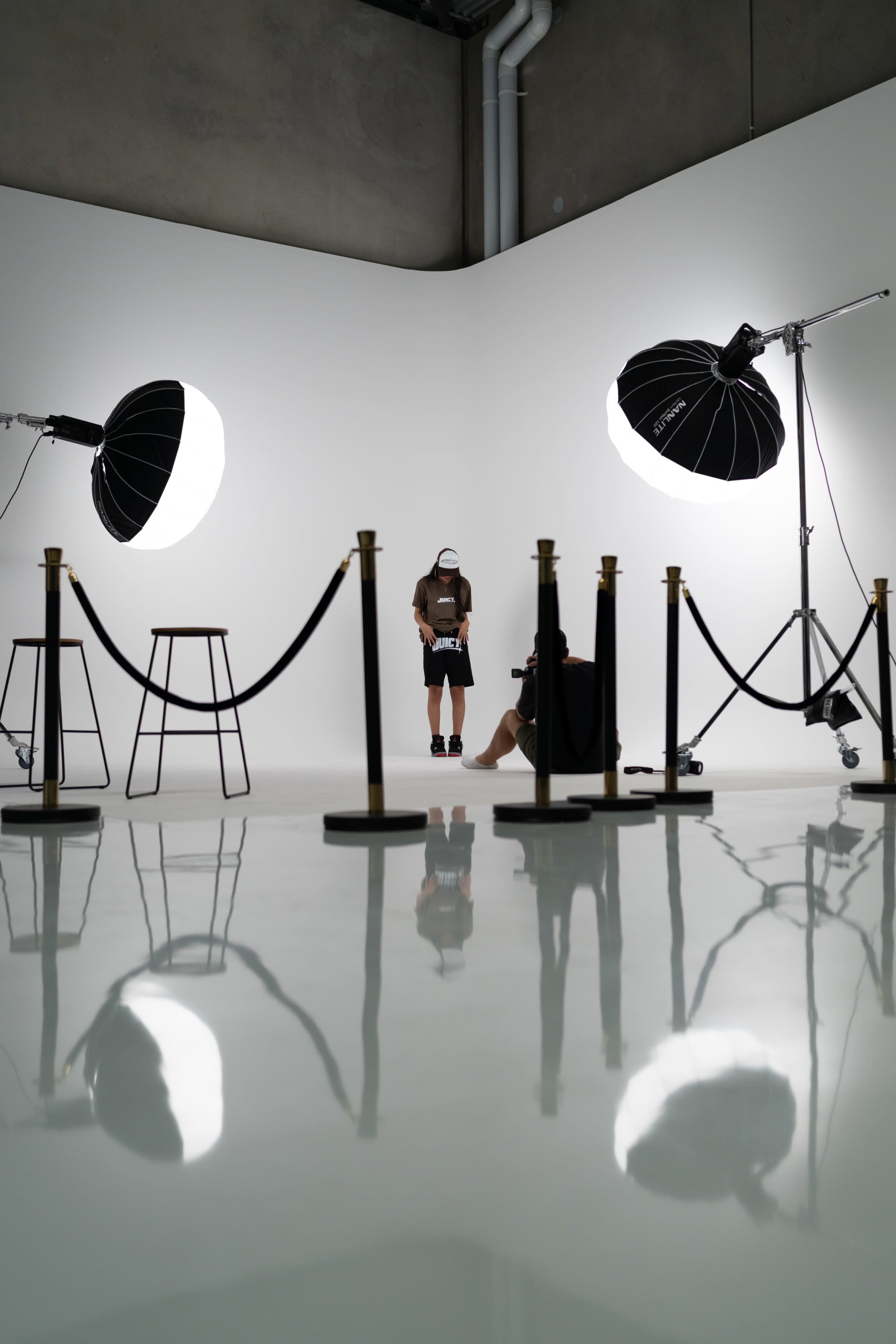 A photoshoot setup features a model and a photographer in a studio with two large light sources and a roped-off area.