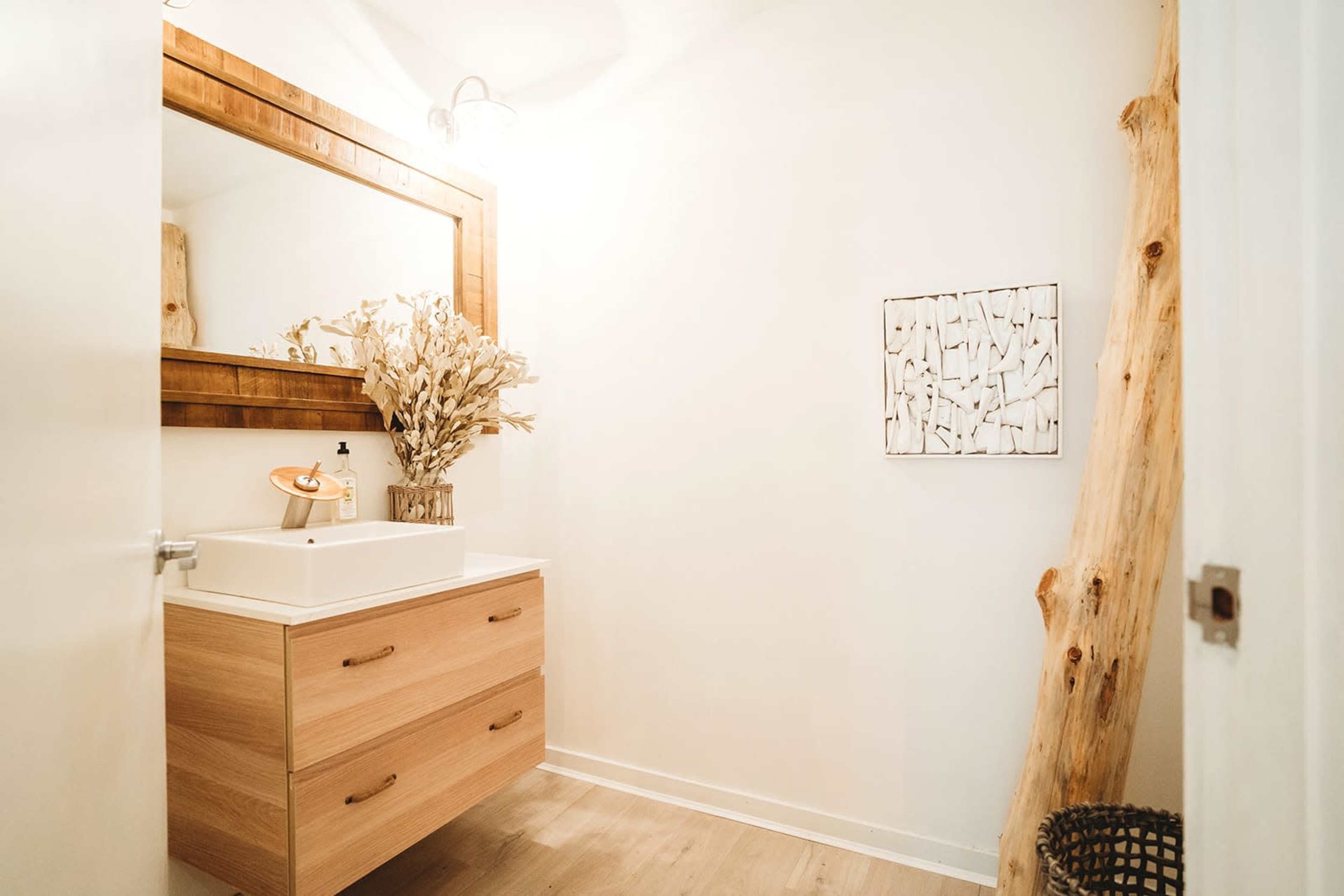 A minimalist bathroom with a wooden vanity, a large mirror, a bouquet of dried flowers, and a decorative wall piece.
