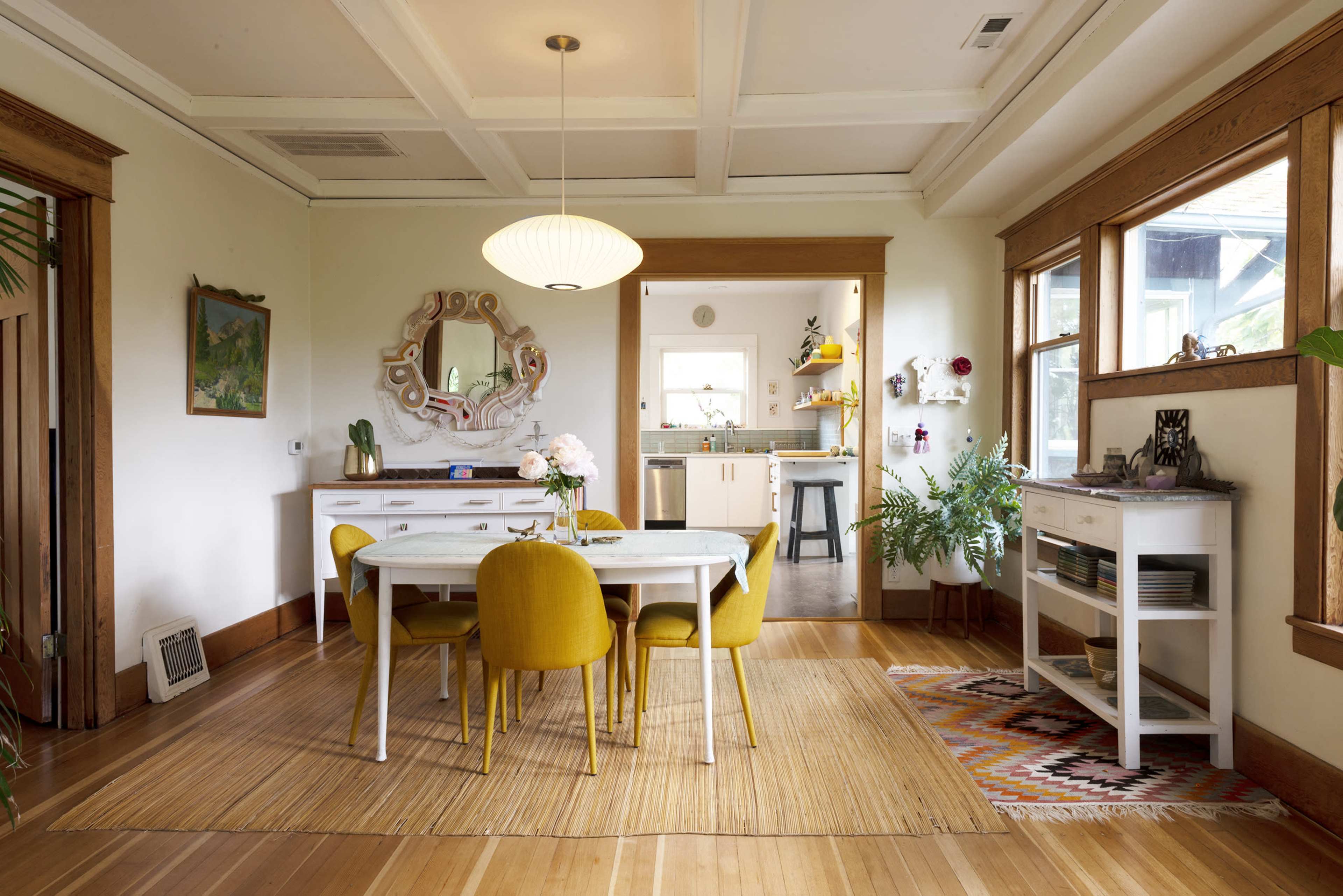 A dining area features a round table surrounded by yellow chairs, with a large mirror and art on the walls, and a view into a kitchen beyond.