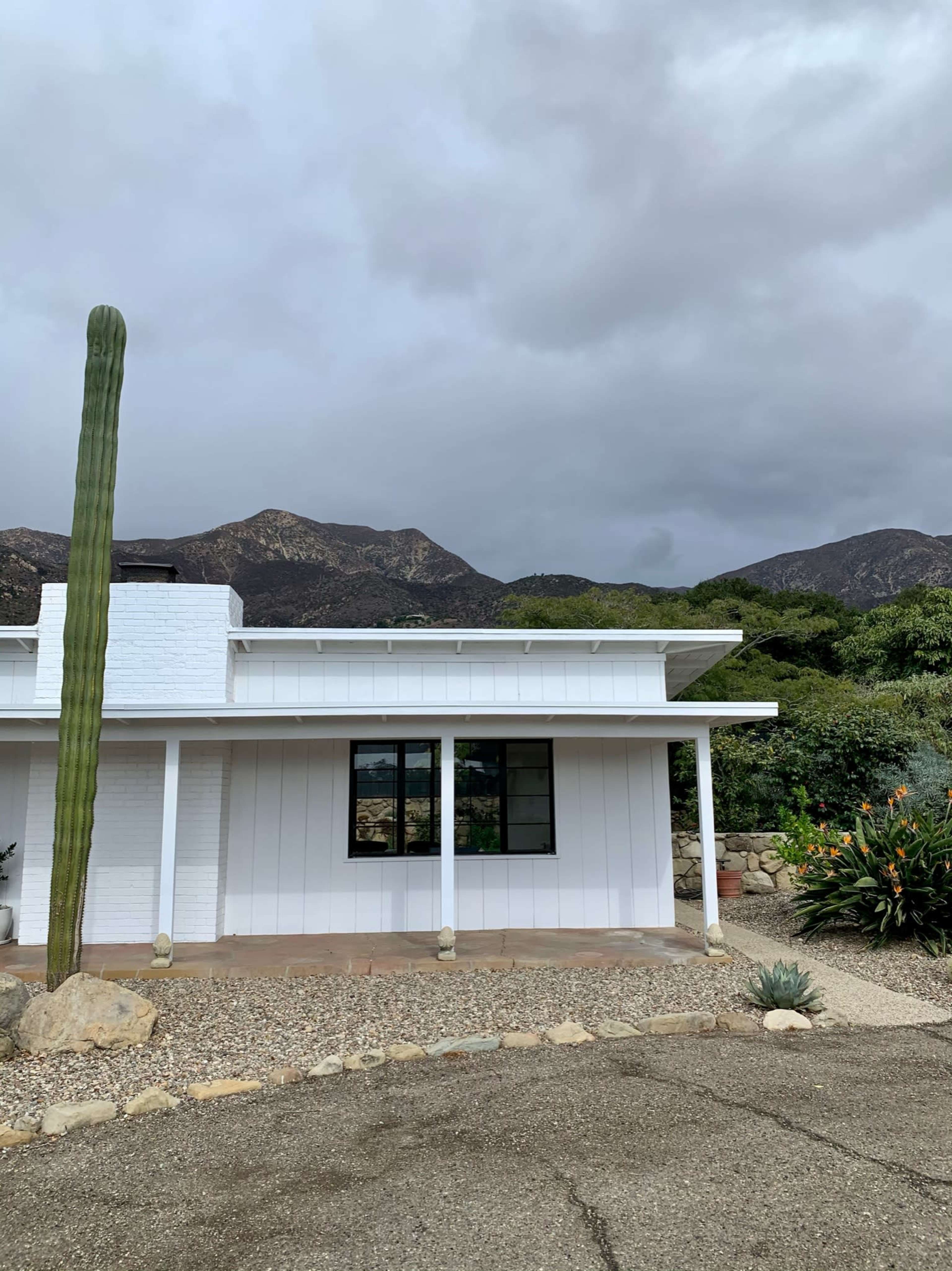 A white house with a large cactus in front is set against a backdrop of mountains and cloudy skies.