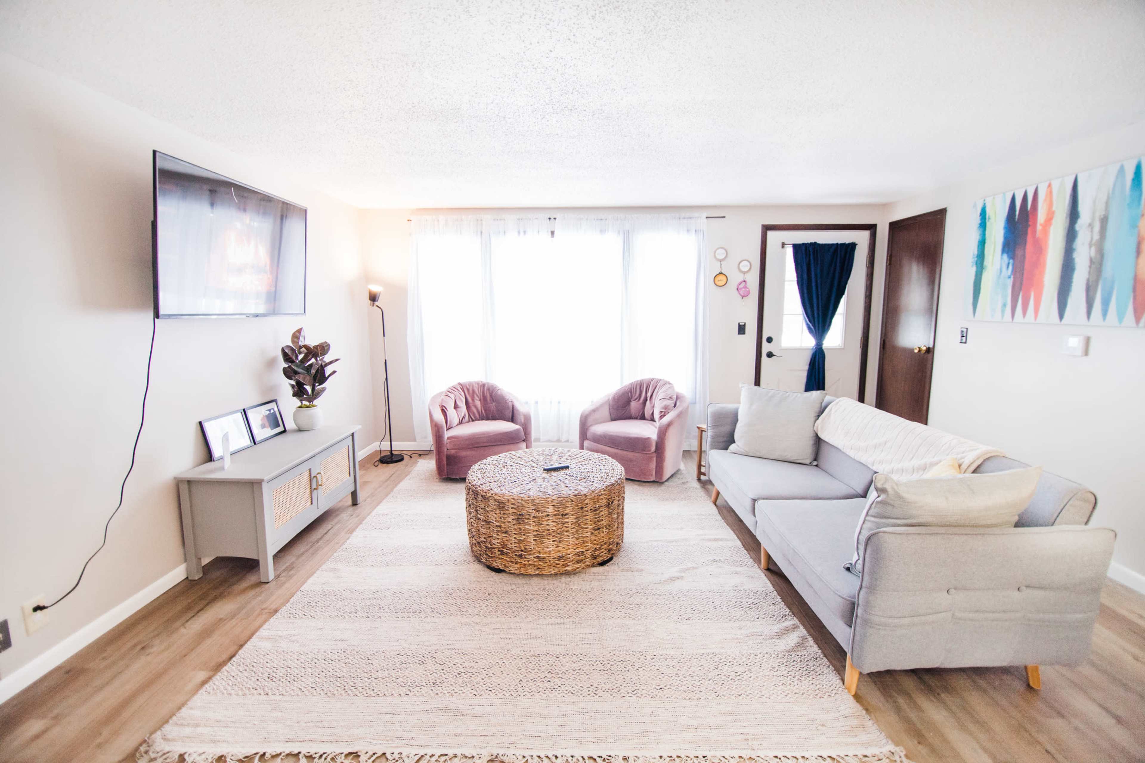 The living room features a gray sofa, two pink armchairs, a round wicker coffee table, and a television on the wall, with a window allowing natural light.