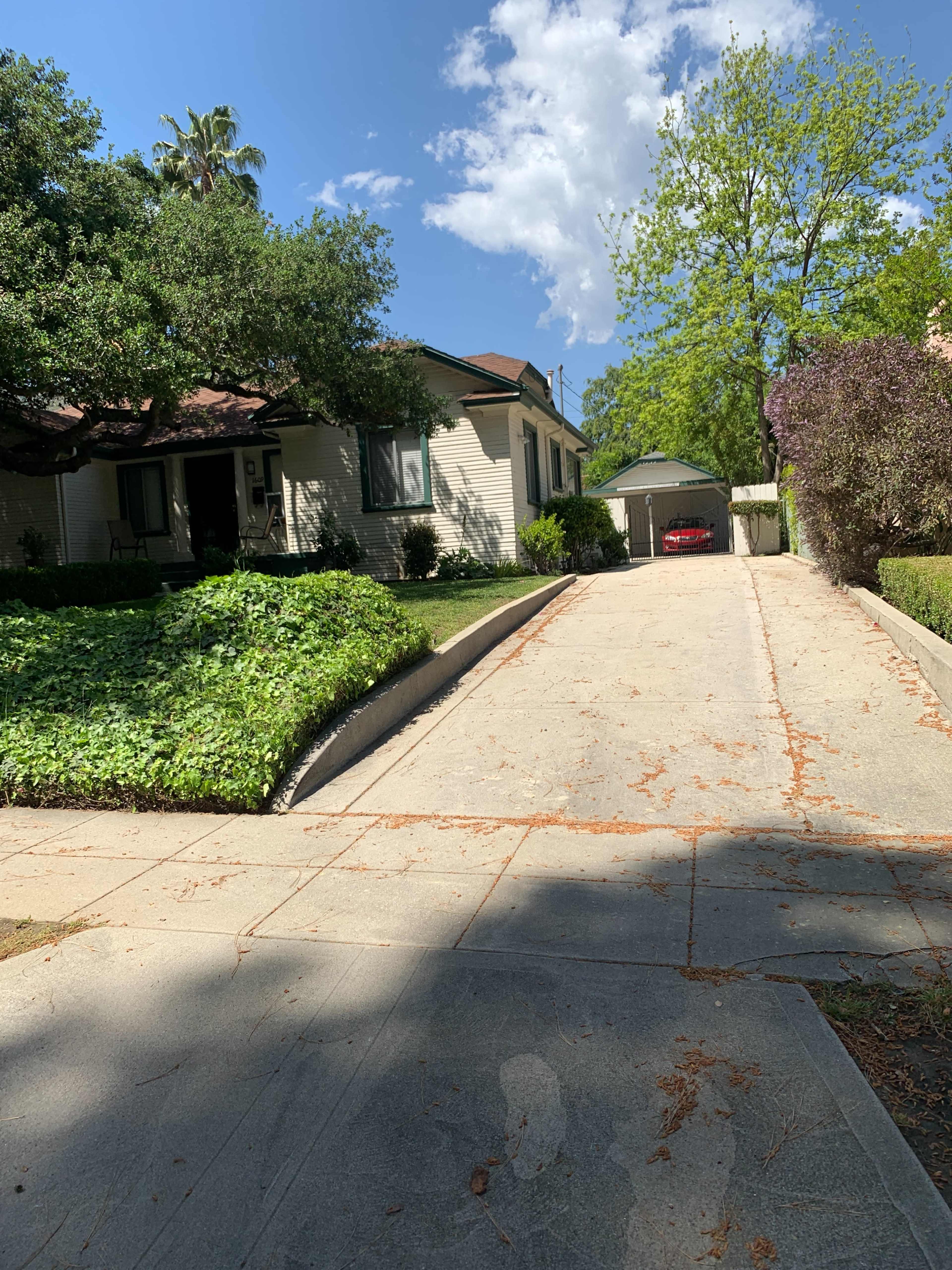 A concrete driveway leads to a house surrounded by green shrubs and trees under a partly cloudy sky.