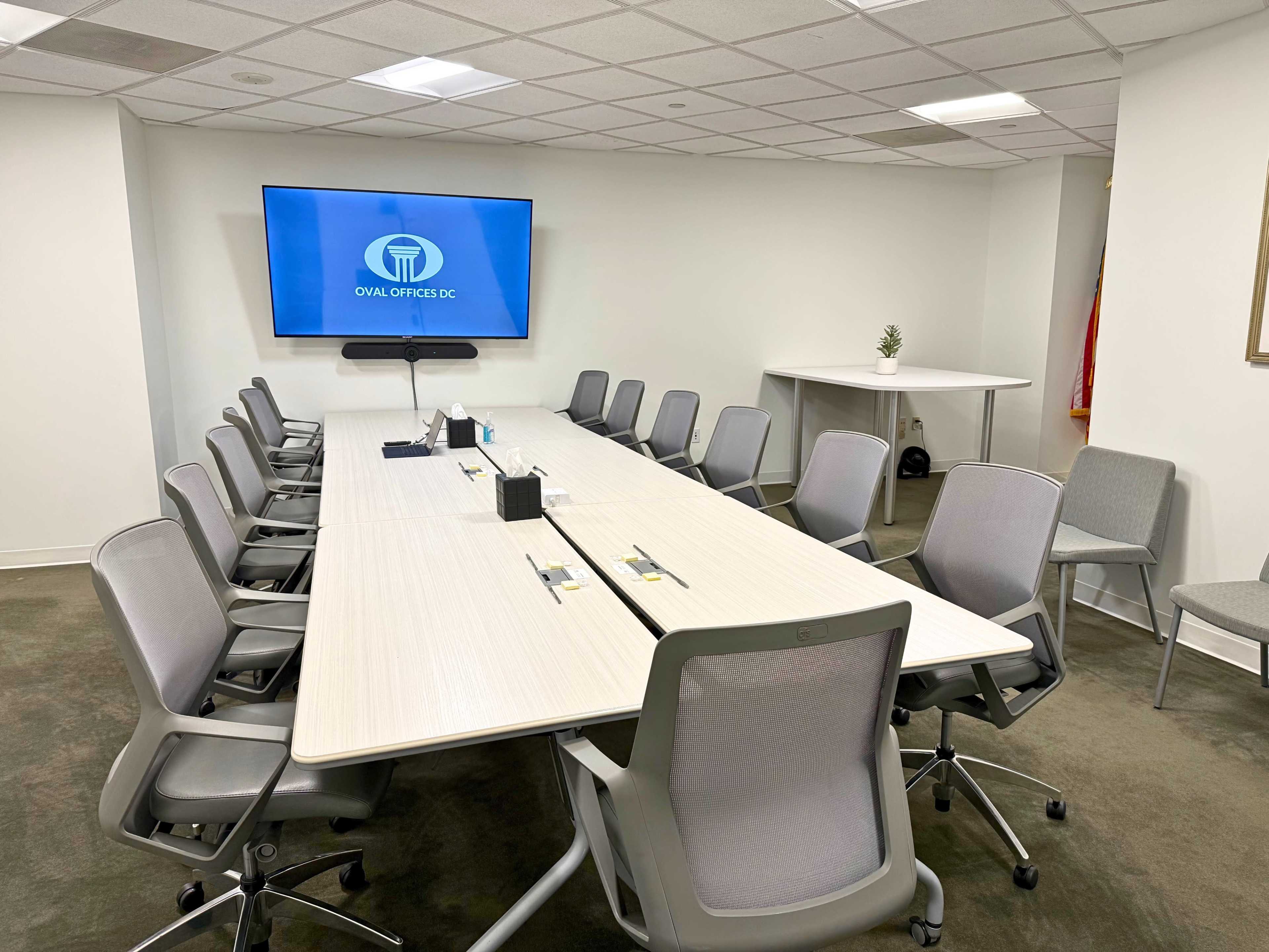 The image shows a modern conference room with a long table surrounded by multiple chairs, and a screen displaying a logo on the wall.