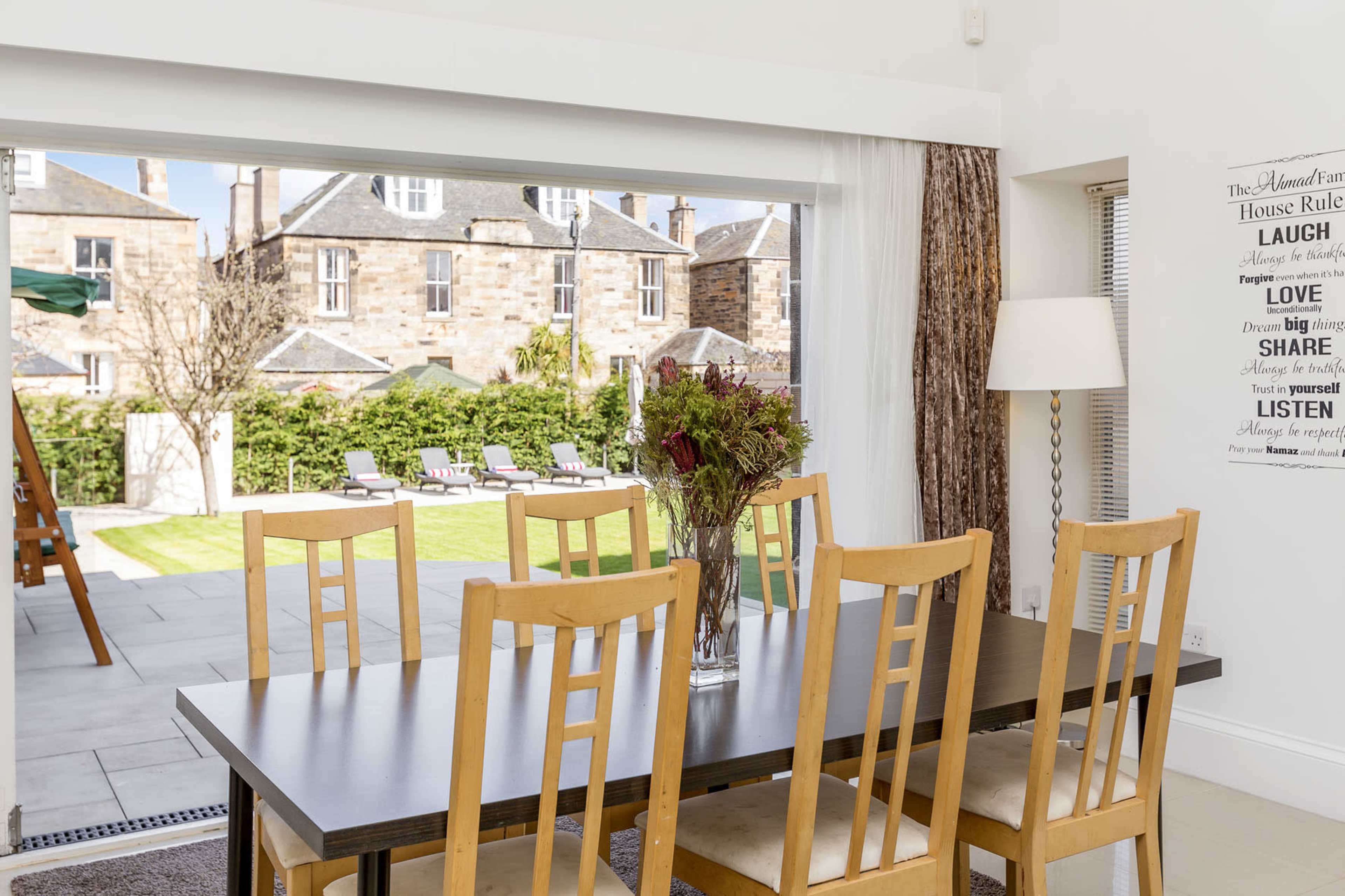 A dining table with wooden chairs is set in a bright room, overlooking a garden with lounge chairs and a building facade in the background.