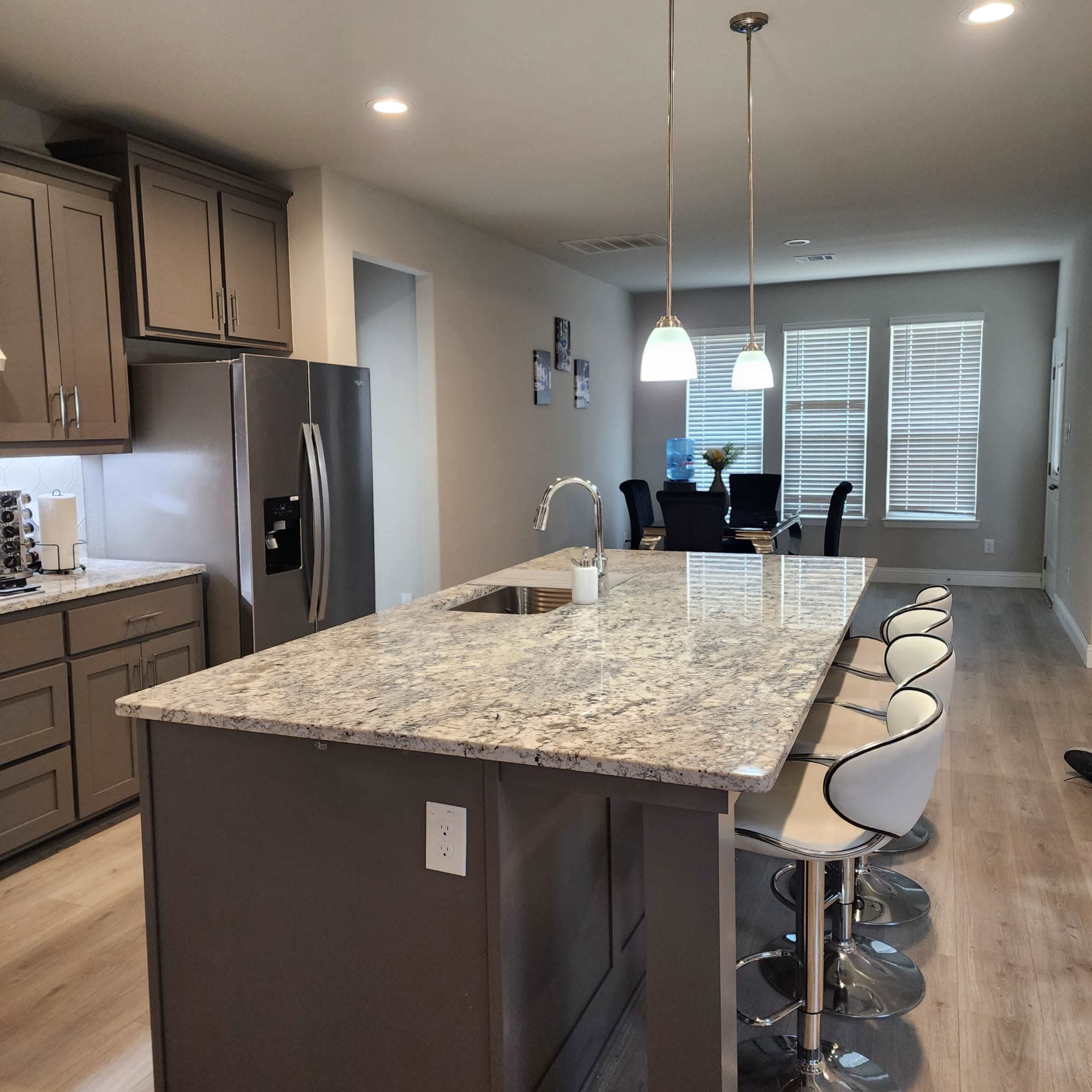 A modern kitchen features gray cabinetry, a granite countertop island with seating, and adjacent dining and living areas with large windows.