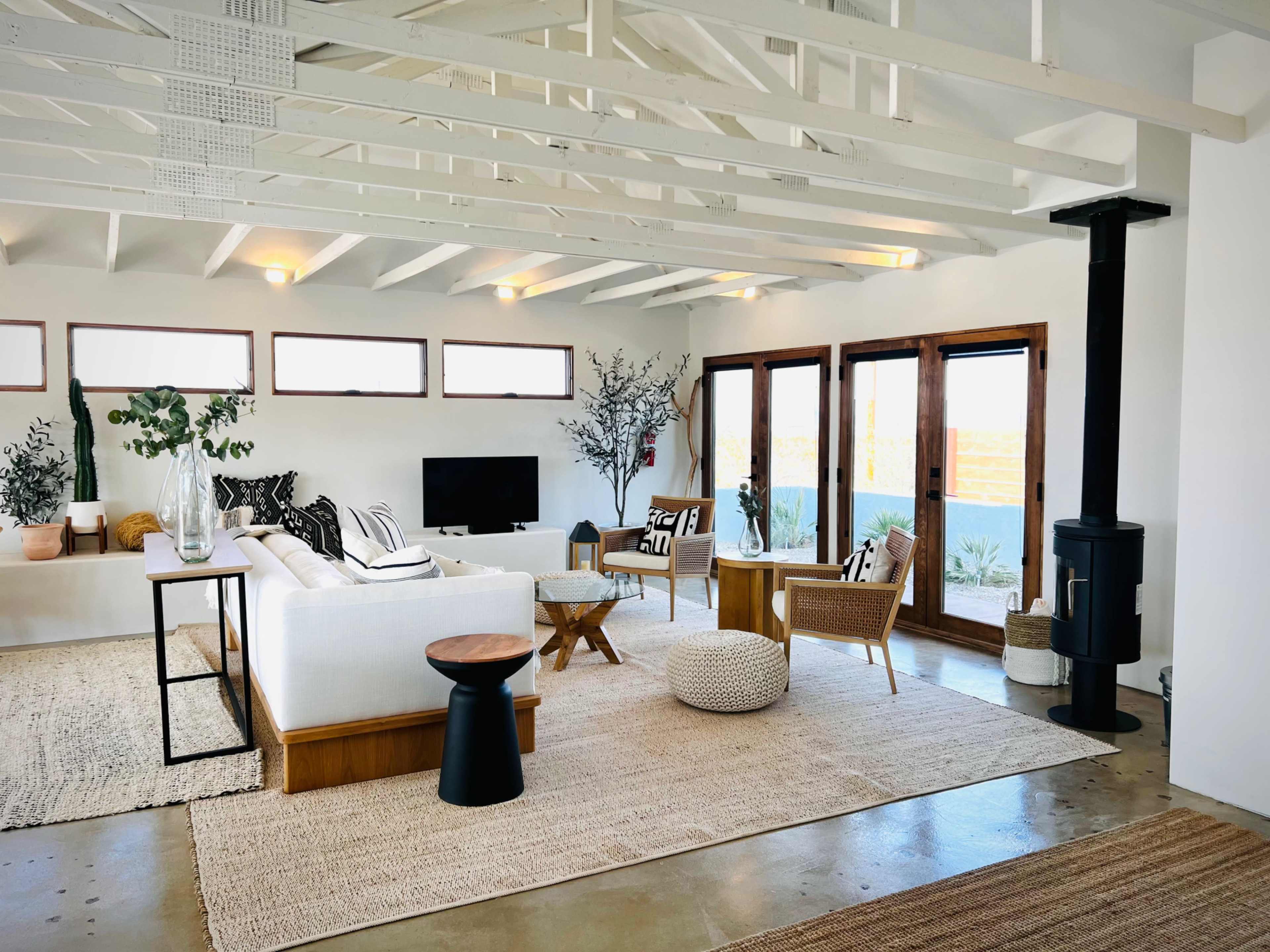 A bright living room with a white sofa, wooden furniture, a rug, and large windows offering a view of the outdoors.