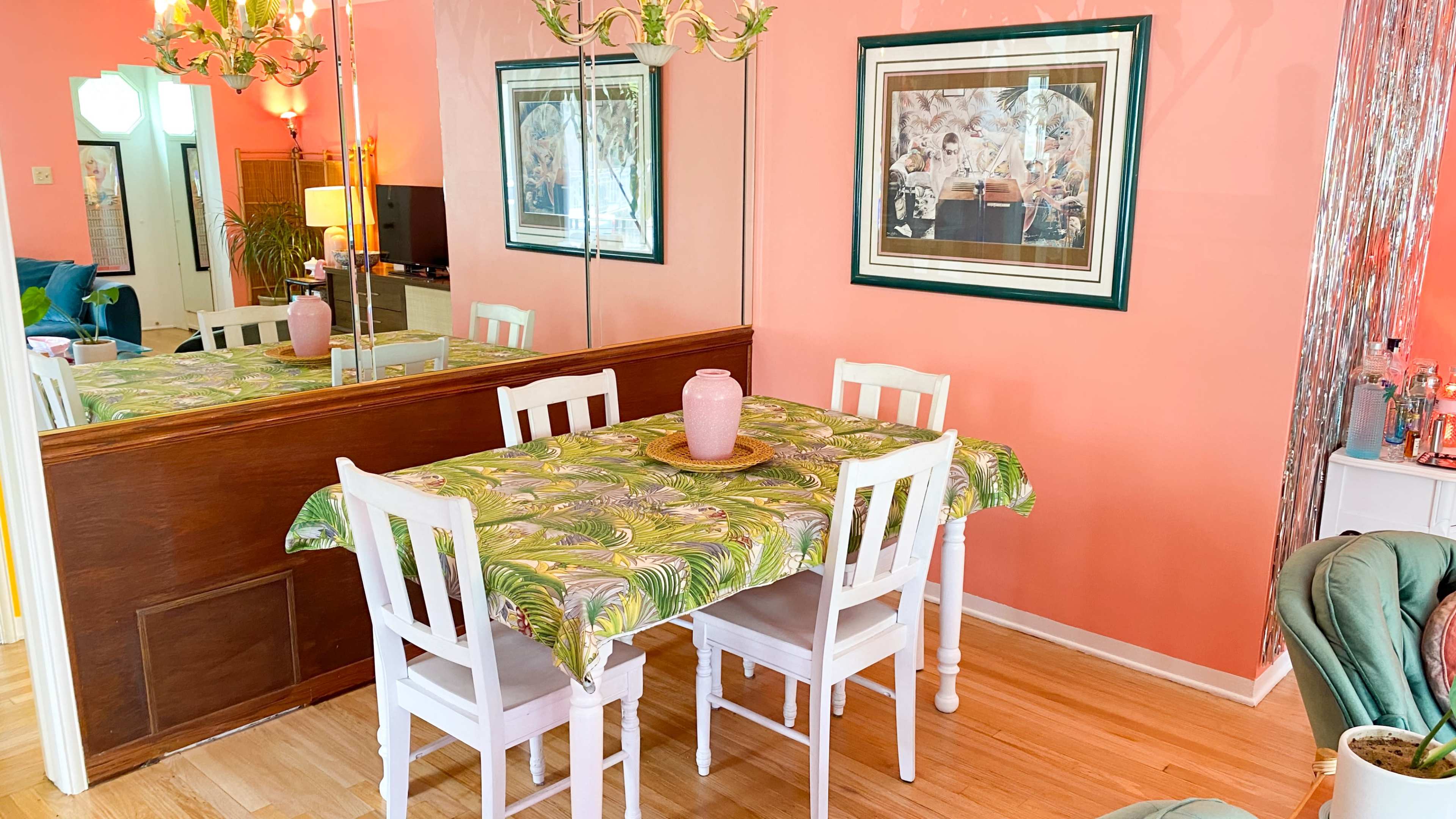 A dining table with a patterned cloth is set in a room with pink walls and a large mirror reflecting the decor.