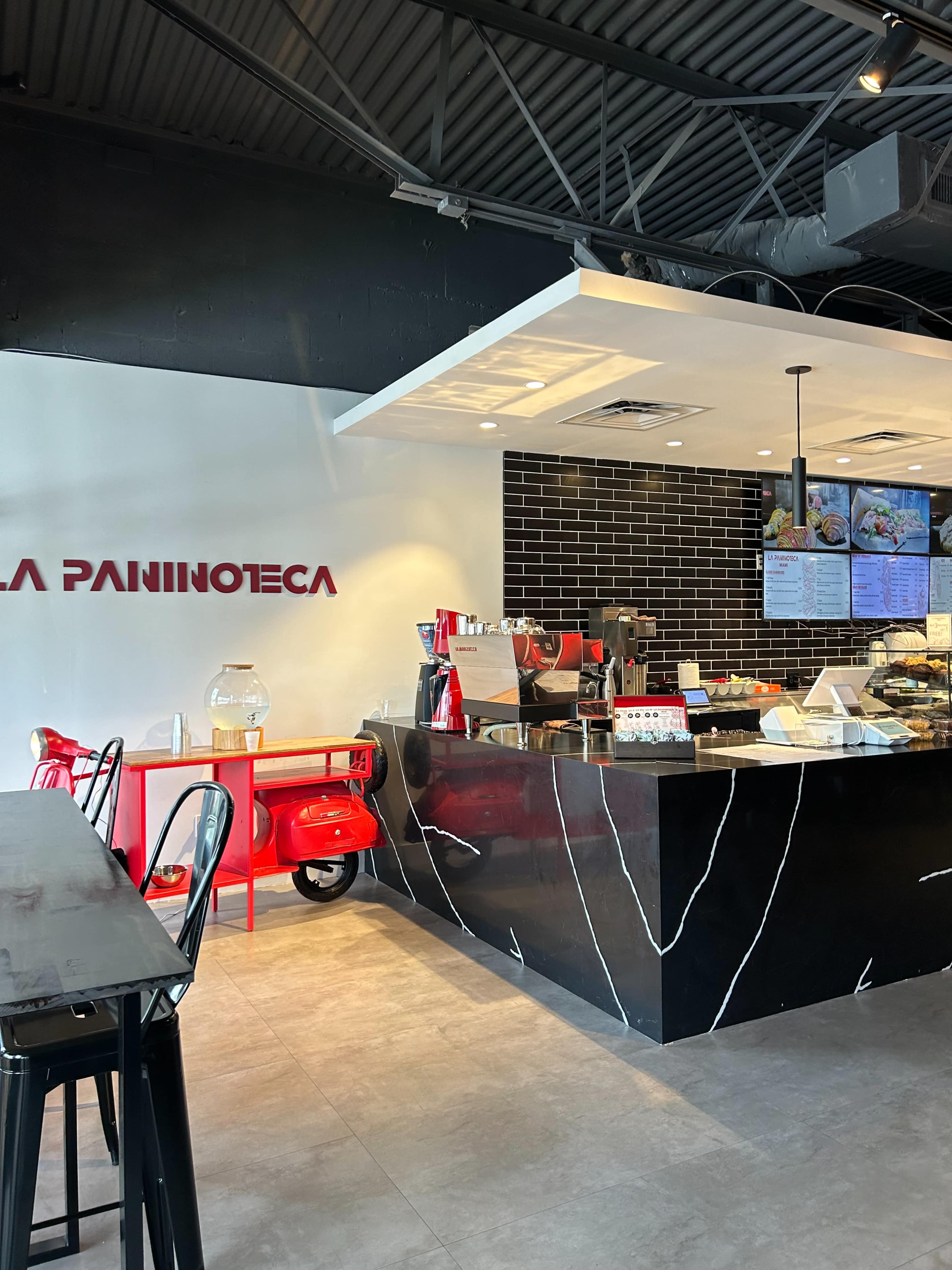 The image shows the interior of a café with a counter featuring a display of food, a coffee machine, and a red scooter parked nearby.