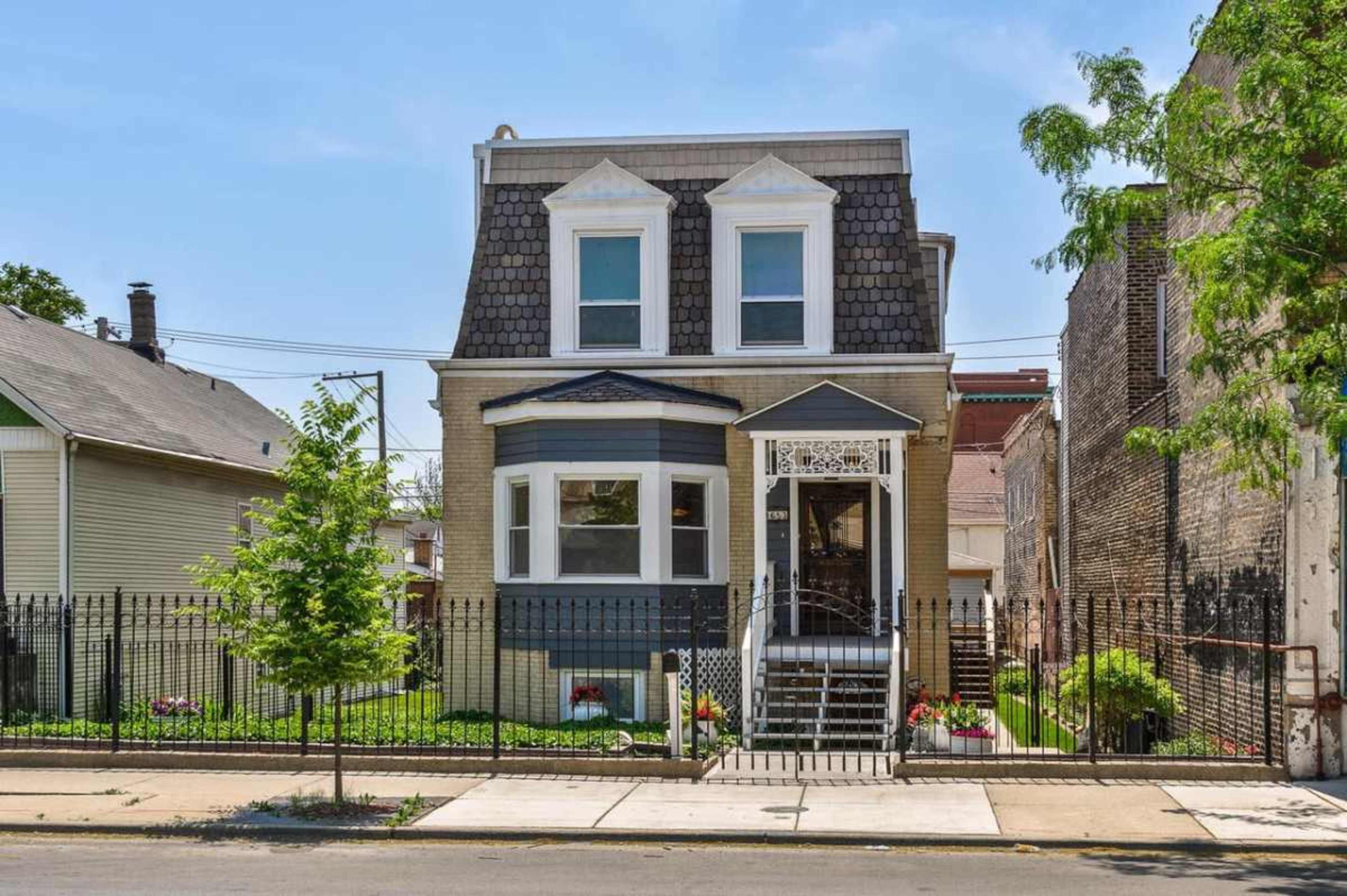 The image shows a two-story house with a gabled roof, surrounded by a fenced yard and a concrete sidewalk.