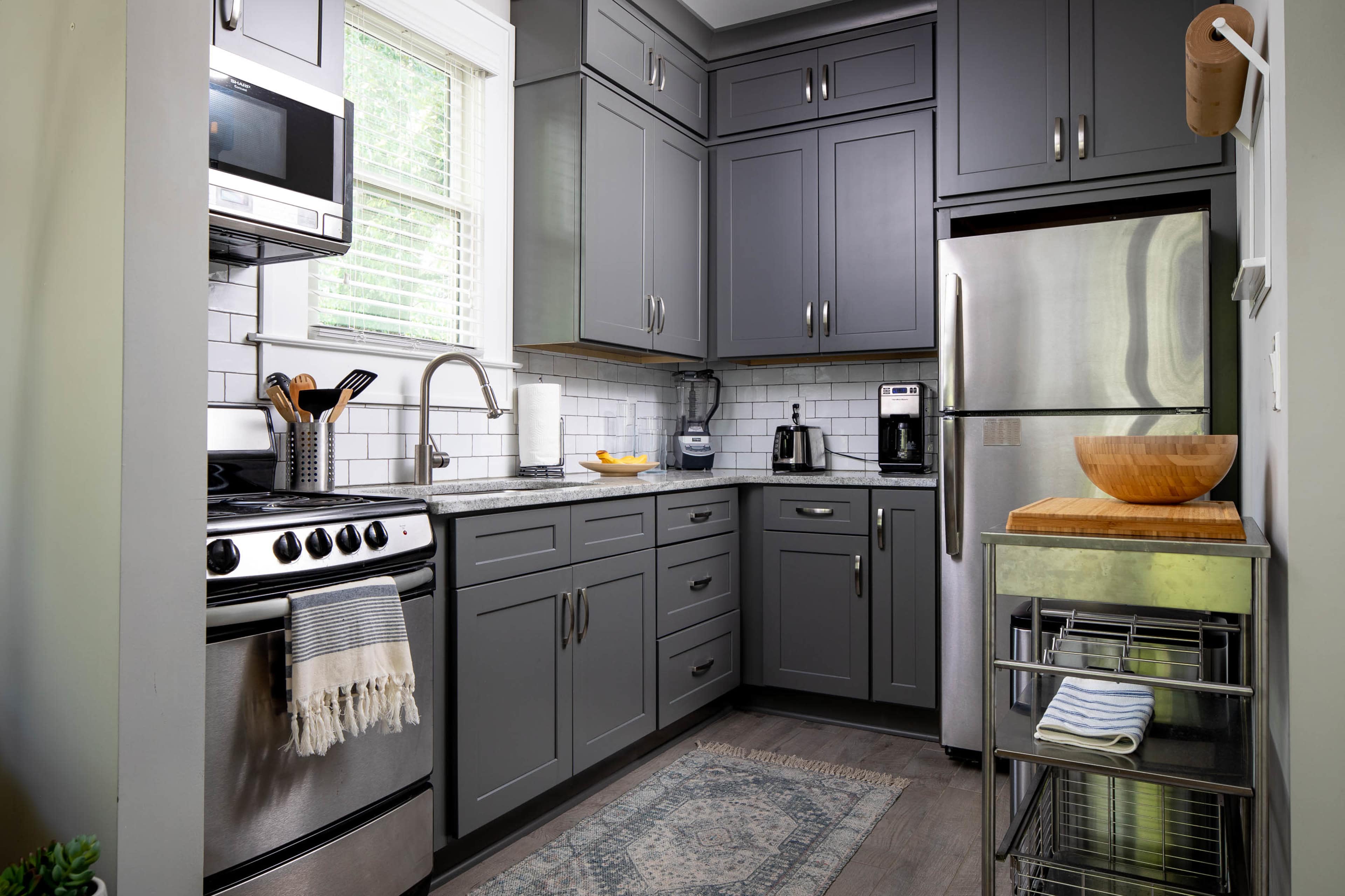 The image shows a modern kitchen with gray cabinets, stainless steel appliances, and a small dining area featuring a wooden bowl and a decorative rug.
