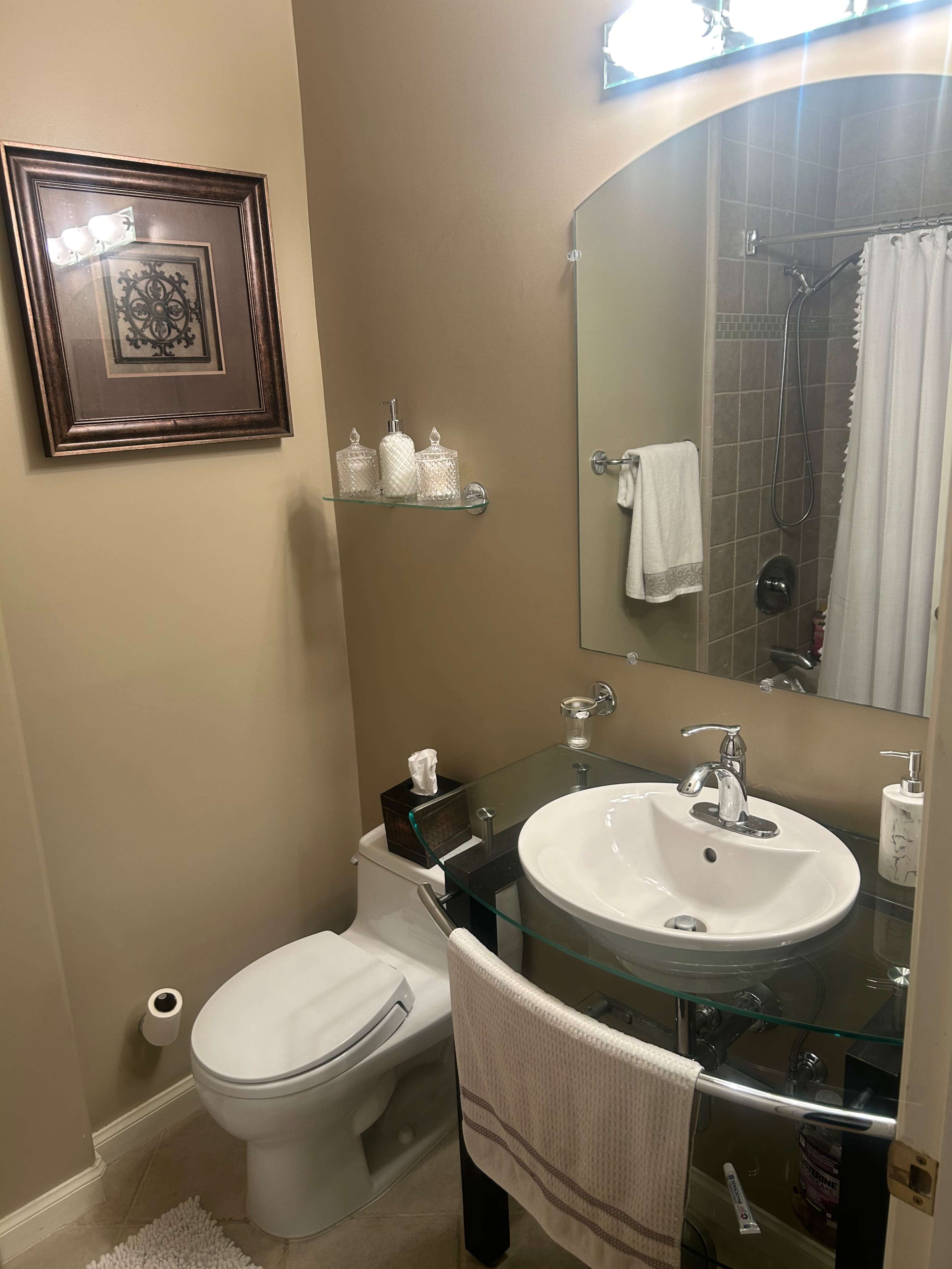 The image shows a small bathroom featuring a glass vanity with a round sink, a toilet, a mirror, and tiled walls.