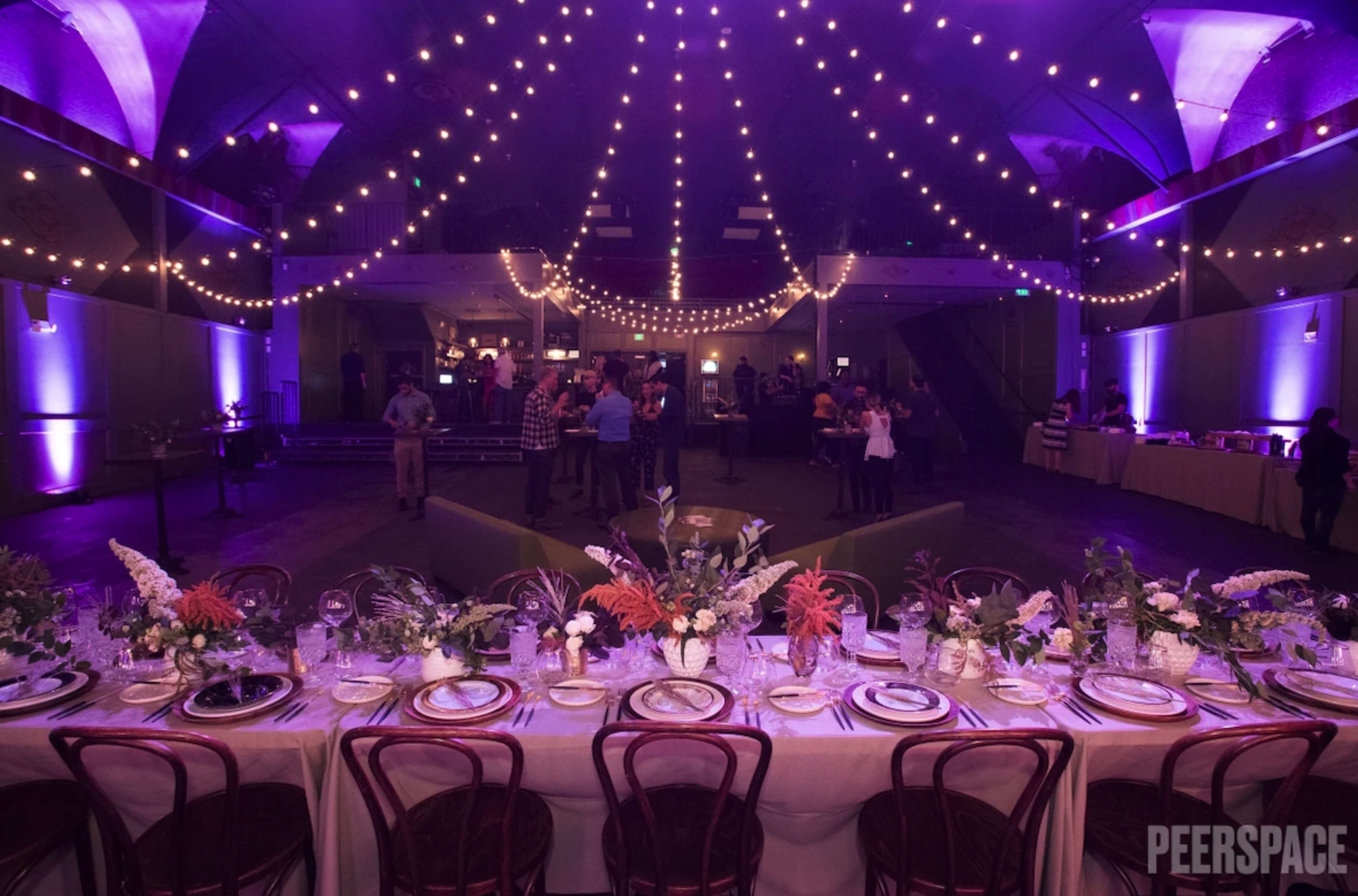 A long dining table is set with floral centerpieces and elegant dinnerware under a canopy of string lights in a warmly lit event space.