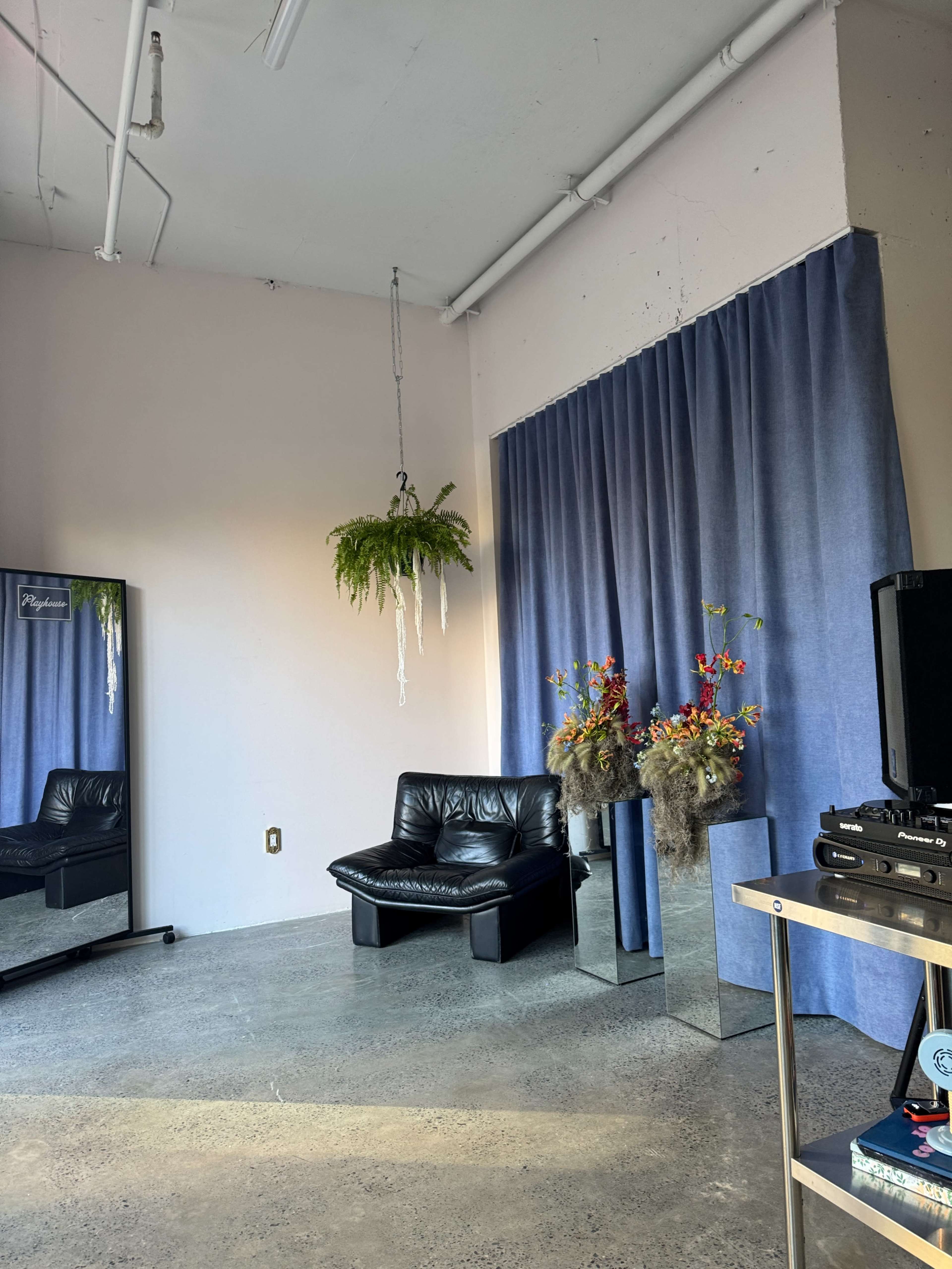 The image shows a minimalist interior space featuring a black leather couch, a large mirror, a decorative potted plant hanging from the ceiling, and a corner with flowers arranged in reflective vases.