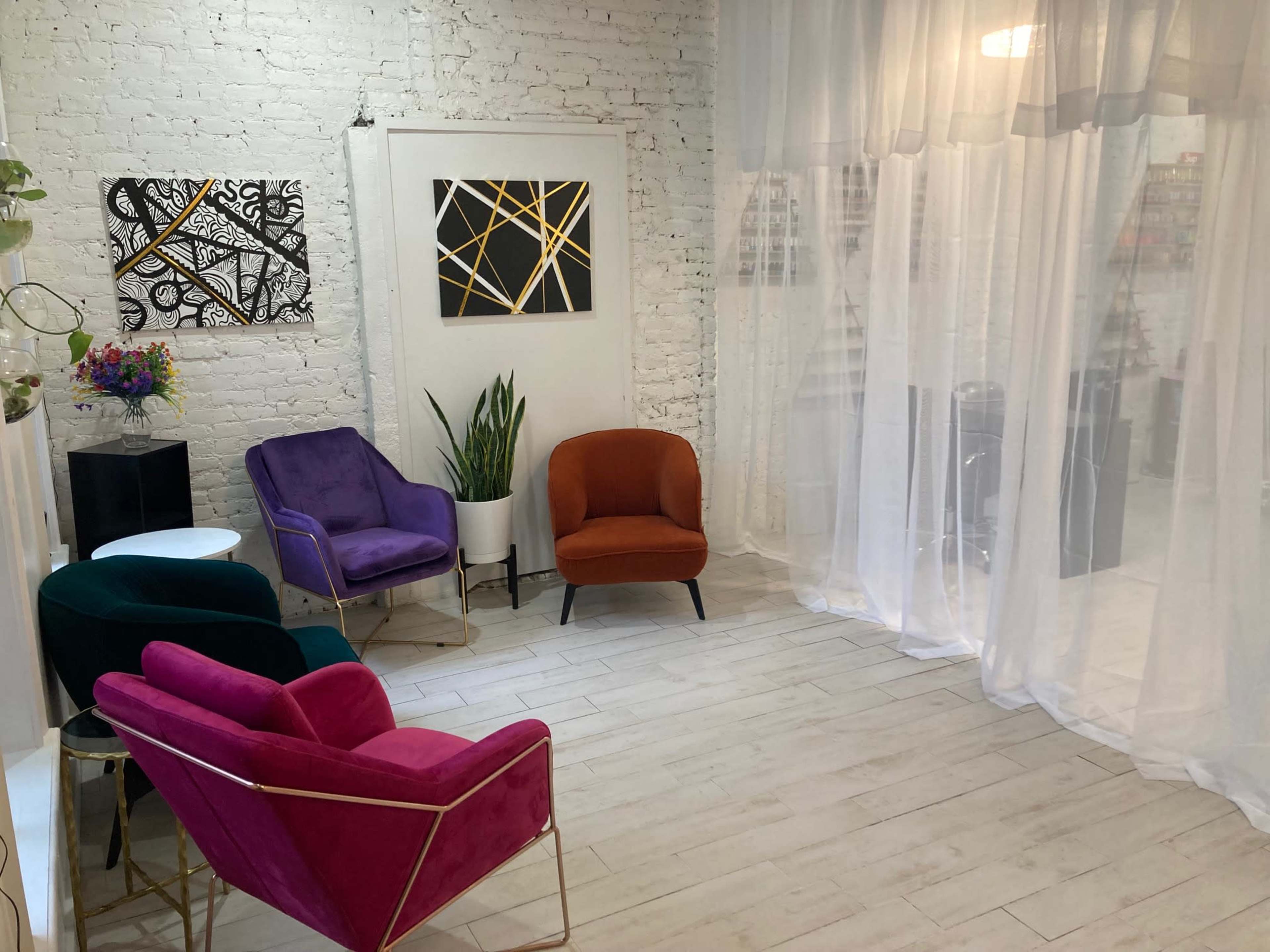 The image shows a bright, minimalist room with four colorful velvet chairs arranged around a small table and abstract art on the walls.