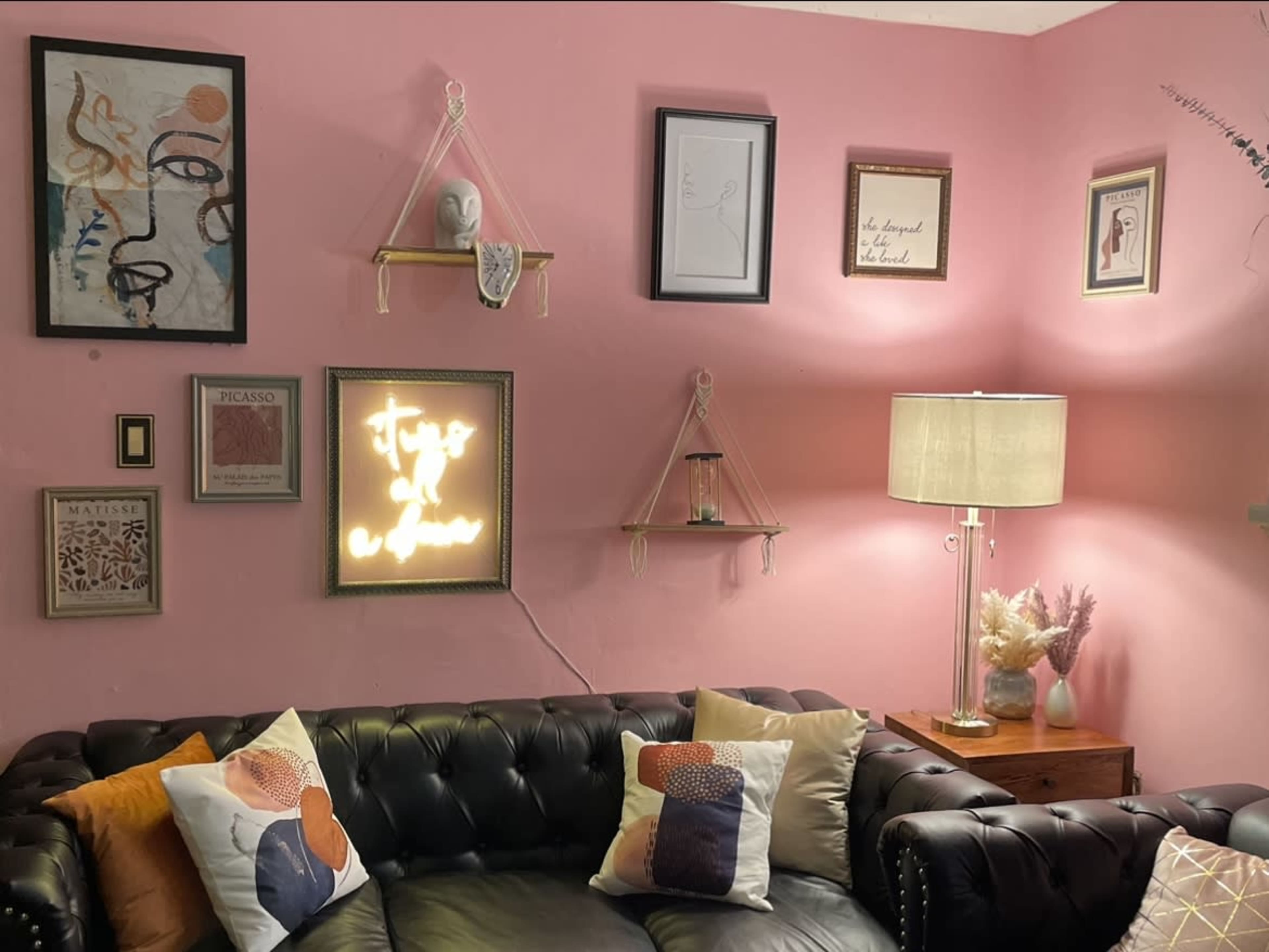 A pink wall adorned with framed art and shelves, alongside a black leather sofa and a lamp.