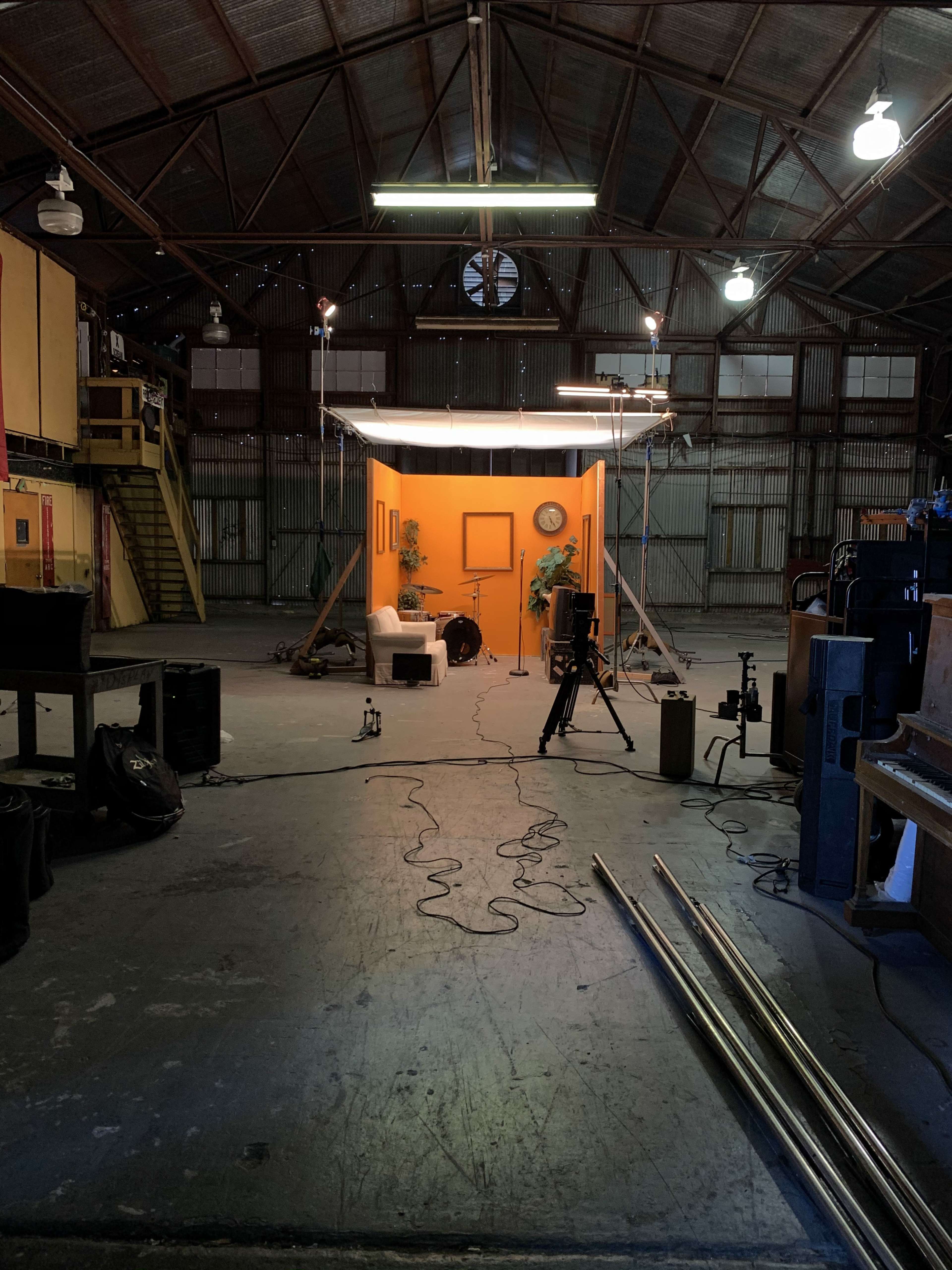 An empty industrial space features a backdrop of orange, with a small sofa, plants, and cameras set up for a photoshoot.
