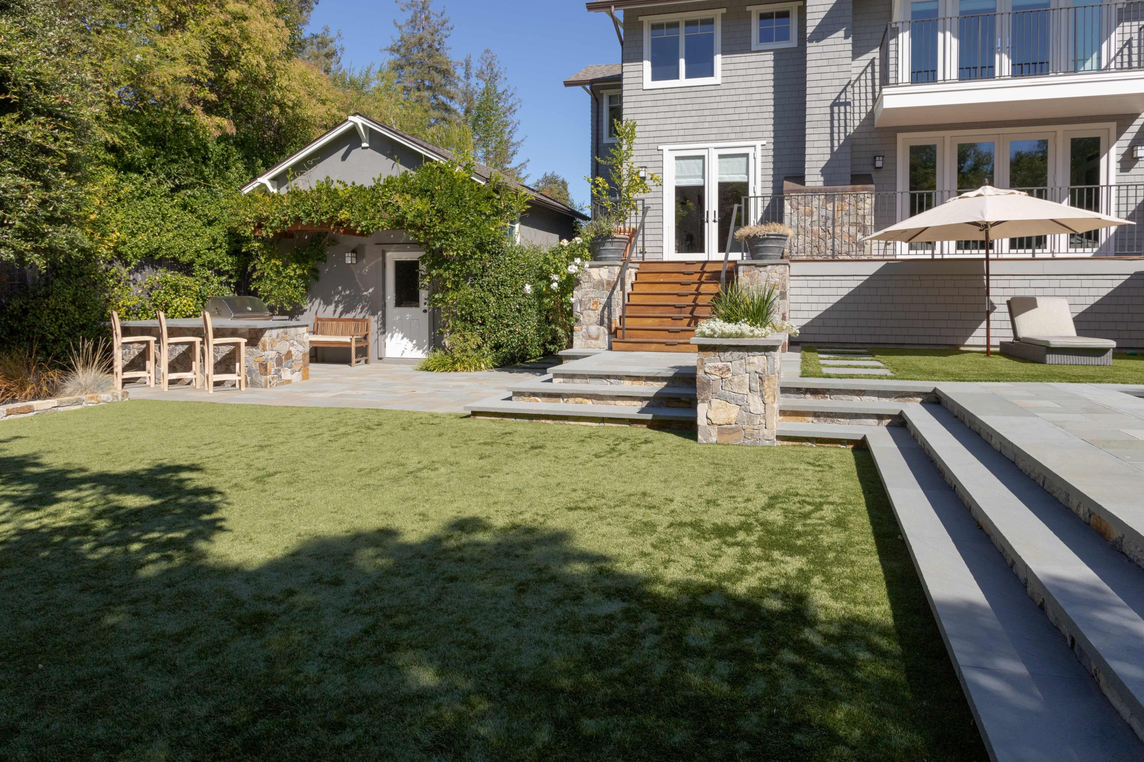 A landscaped backyard featuring a green lawn, stone steps leading to a modern house, and a patio with an umbrella.