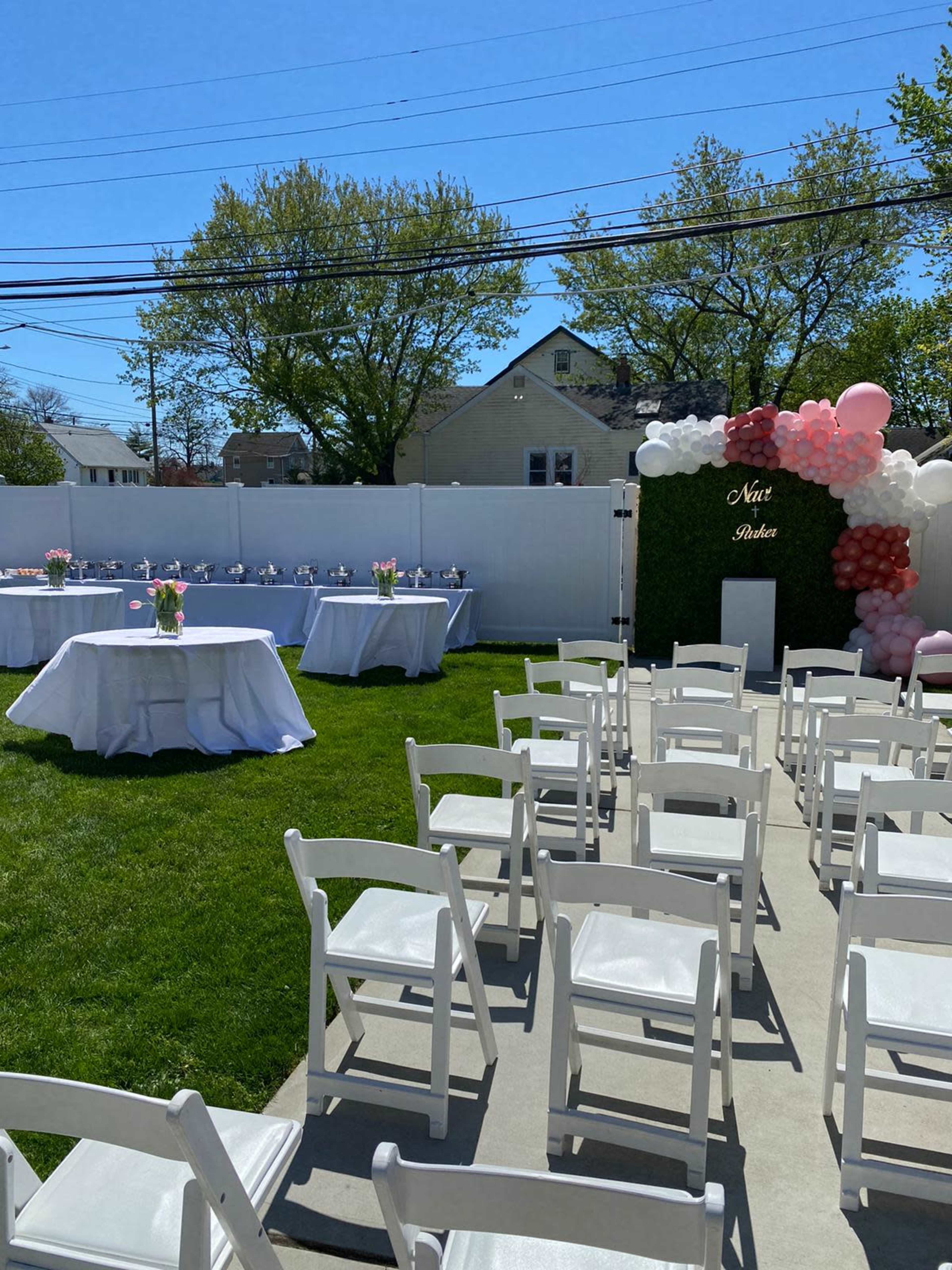 A lawn is set up with white chairs arranged facing a table decorated with flowers, alongside a wall adorned with balloons for a celebration.