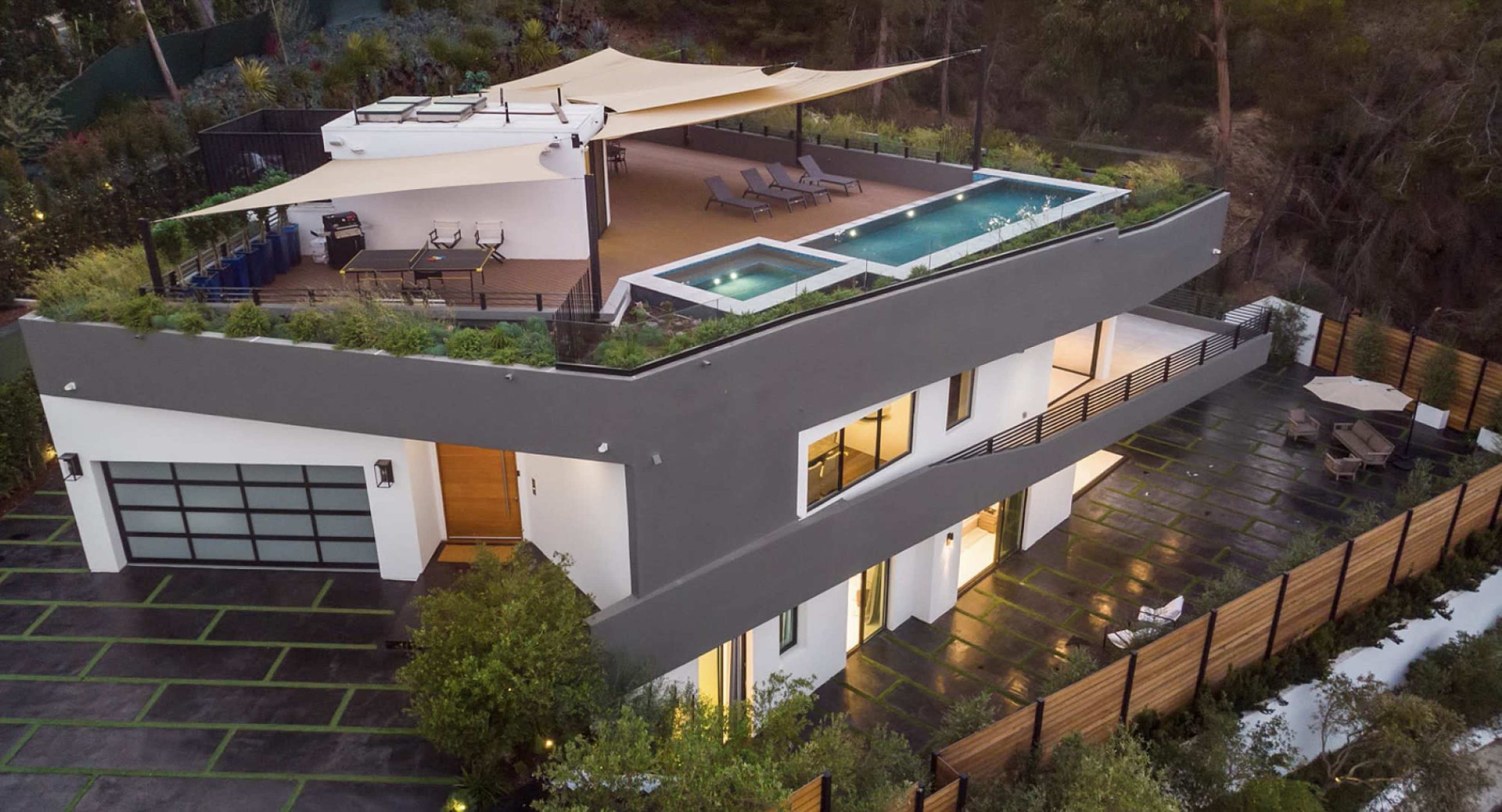 A modern two-story house features a rooftop deck with a pool and seating area, surrounded by greenery and a paved driveway.