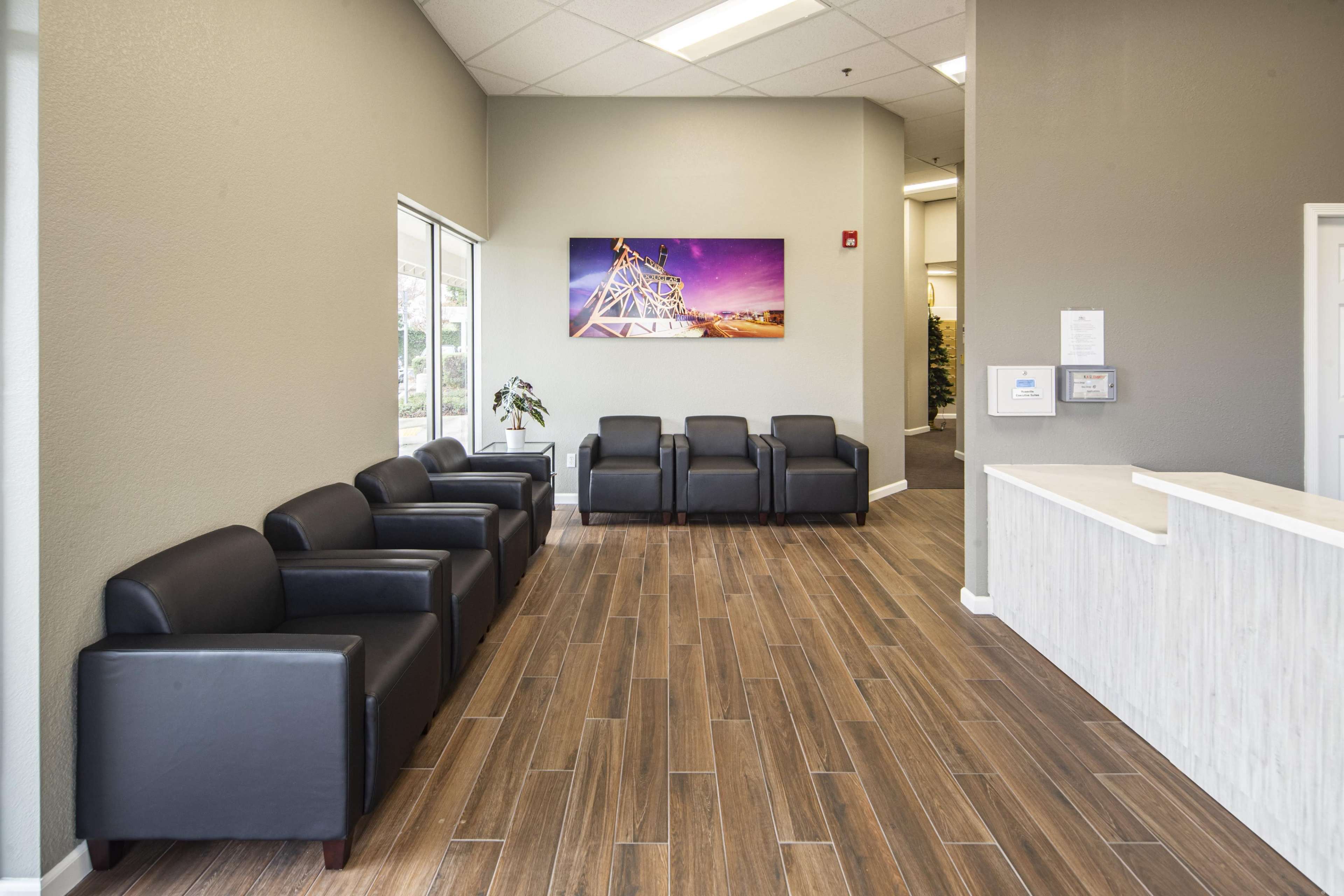 The image shows a modern waiting area with black chairs, a wooden floor, and a bright wall with a colorful artwork above a reception desk.