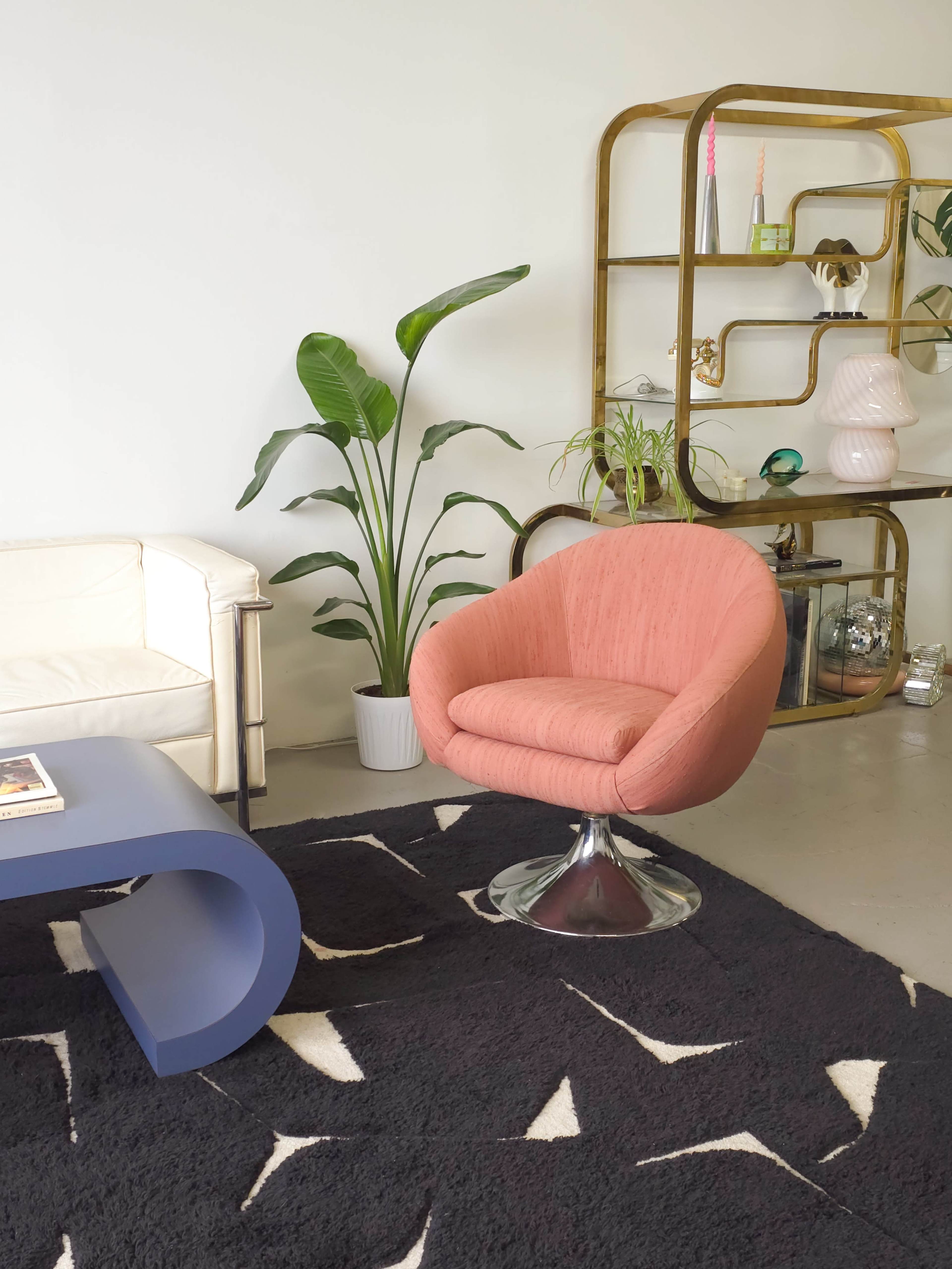 A pink upholstered swivel chair is positioned next to a white leather sofa, with a plant and a gold-framed shelving unit in the background.
