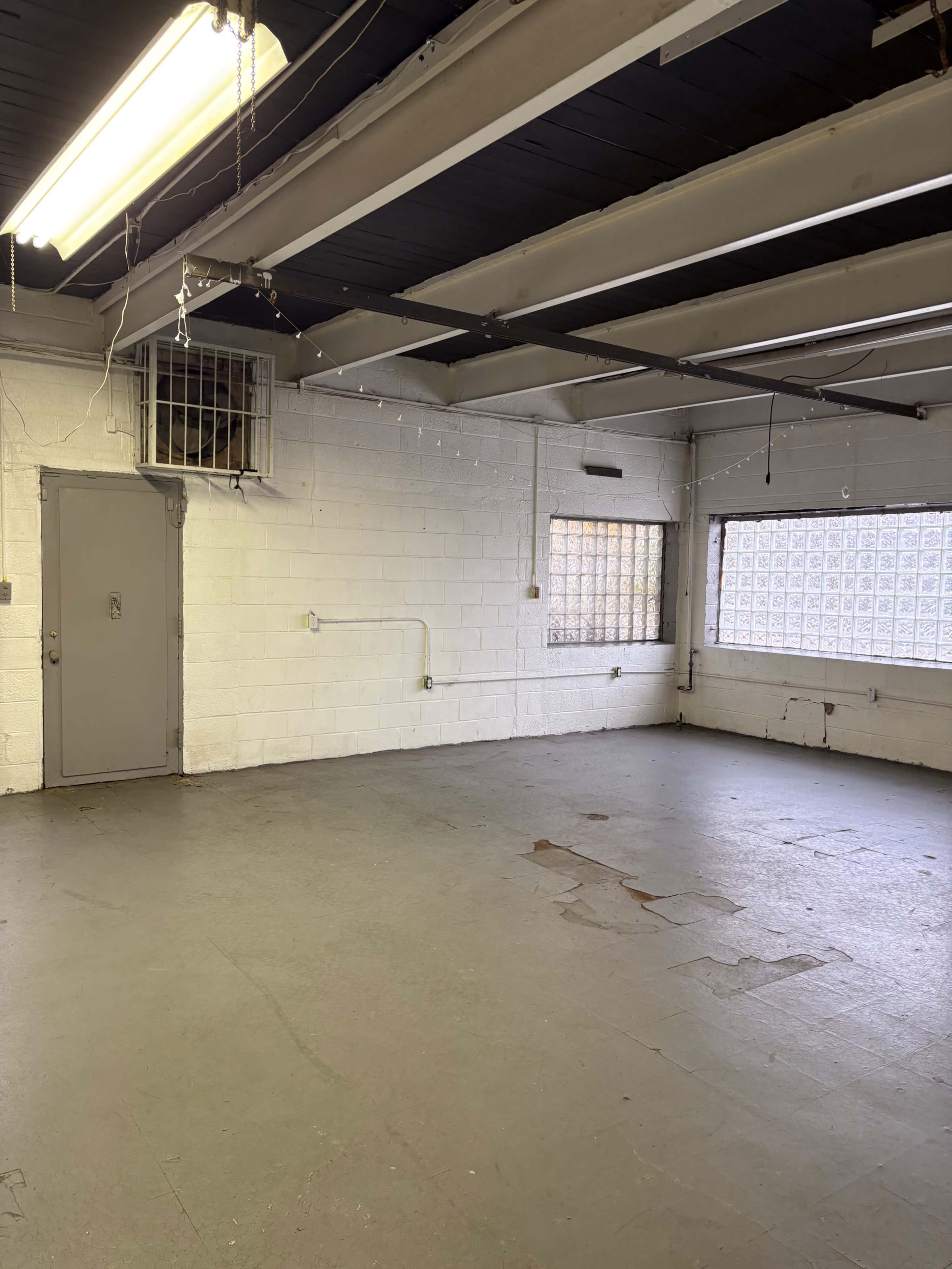 The image shows an empty room with concrete flooring, exposed beams, and two windows featuring glass blocks.