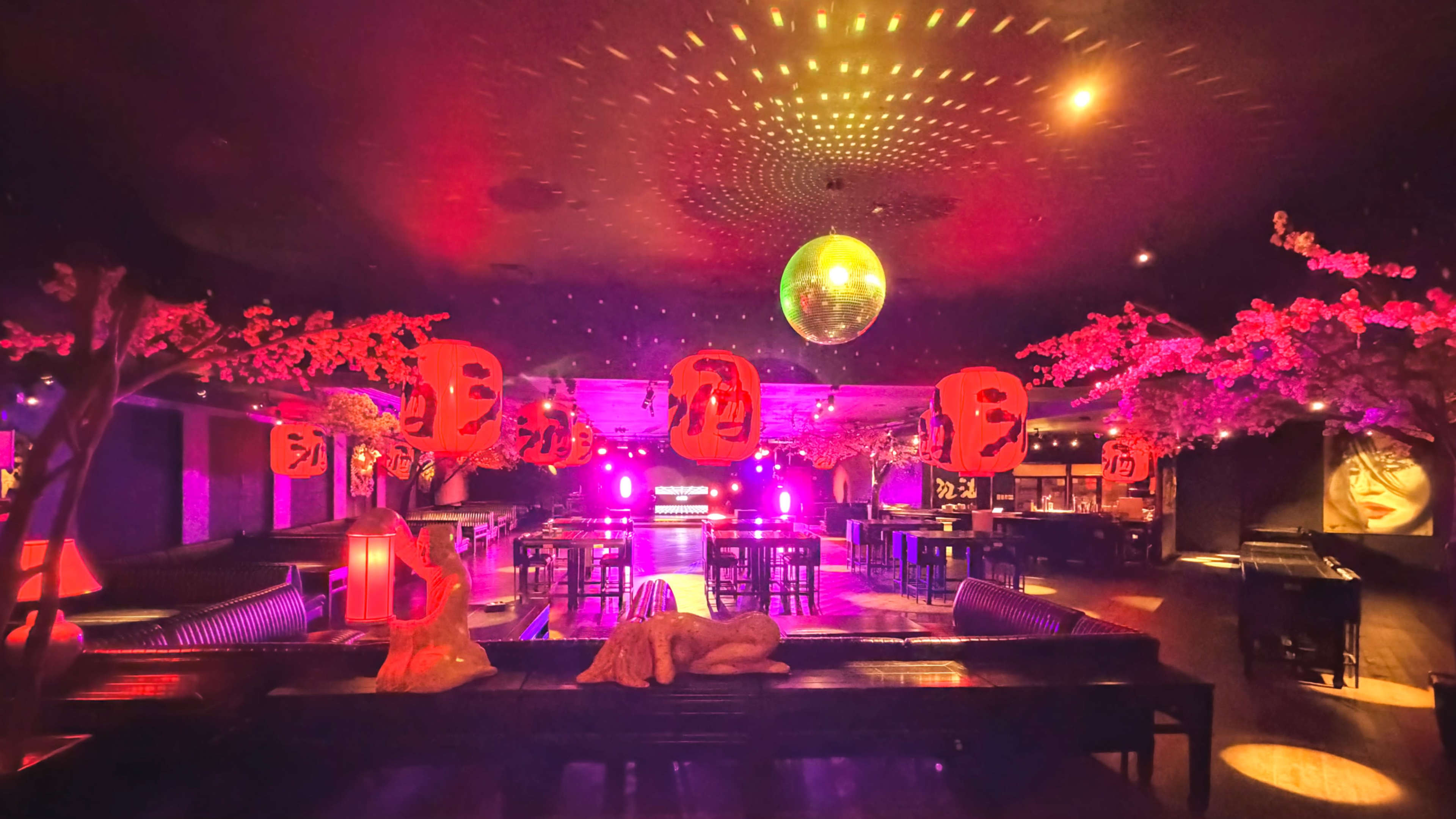 The image shows a dimly lit bar decorated with pink cherry blossoms, red lanterns, and a disco ball, creating a vibrant atmosphere.