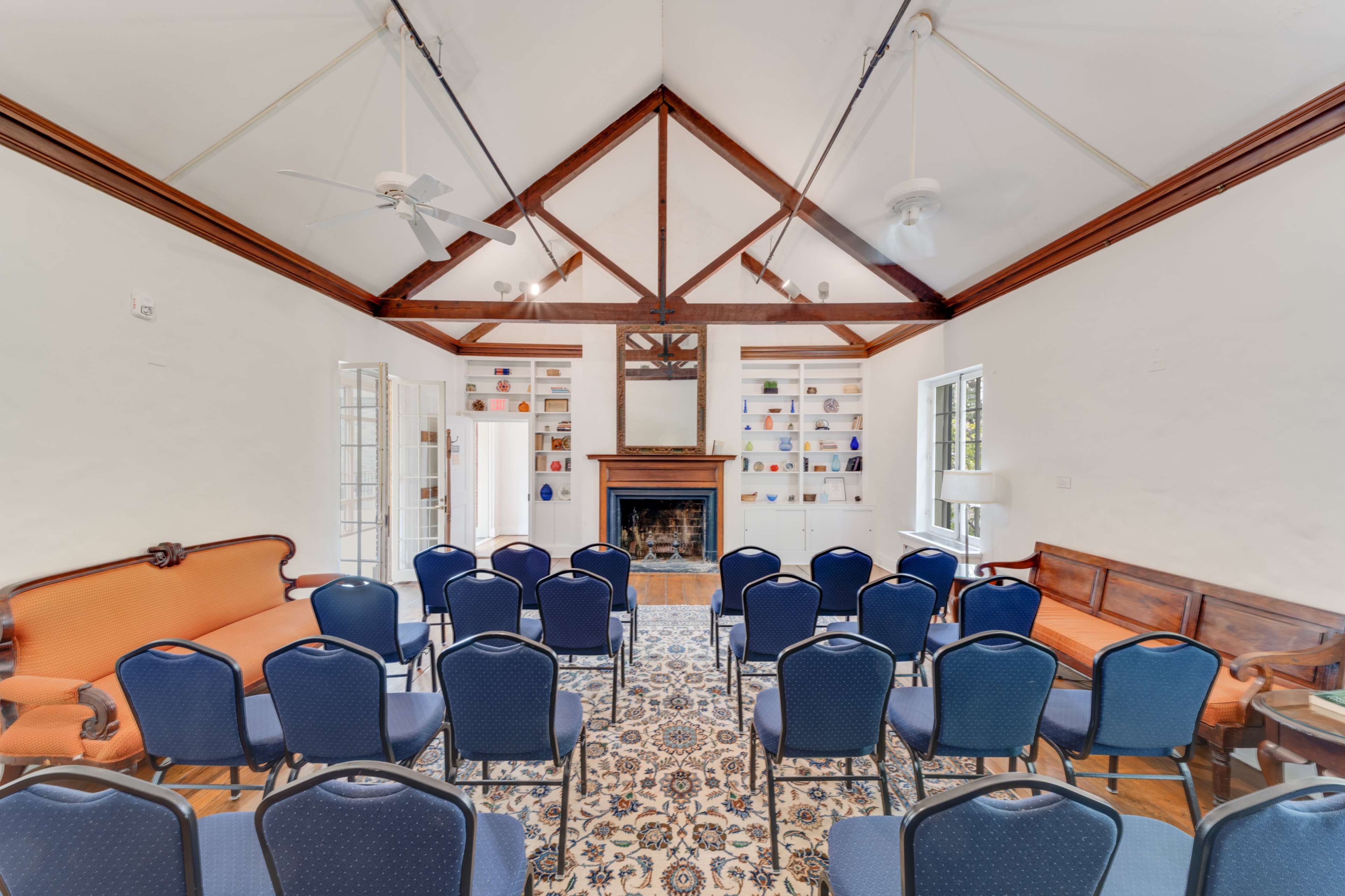 A seminar room arranged with several blue chairs facing a central fireplace, framed by a high ceiling with exposed wooden beams and shelves displaying items on the wall.