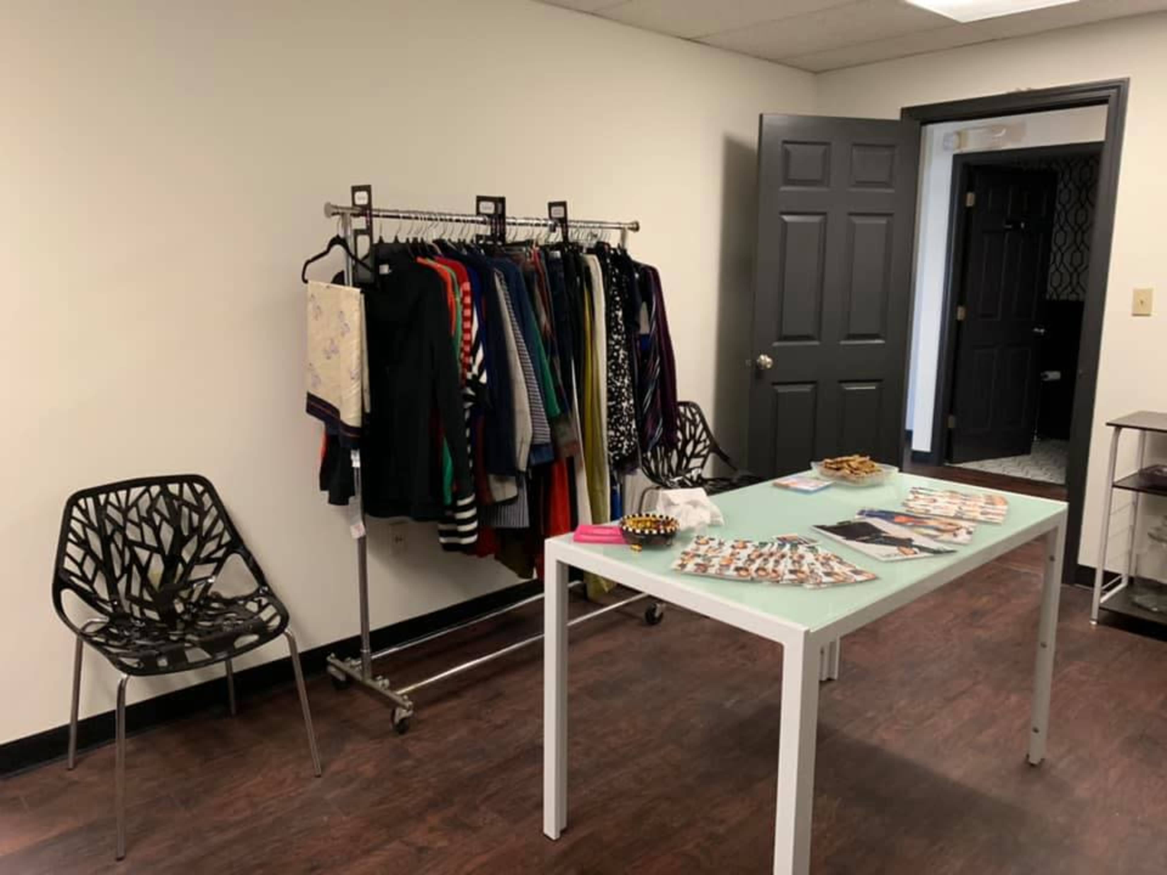 A minimalist room with a clothing rack displaying various garments, a table with accessories and promotional materials, and two black chairs.