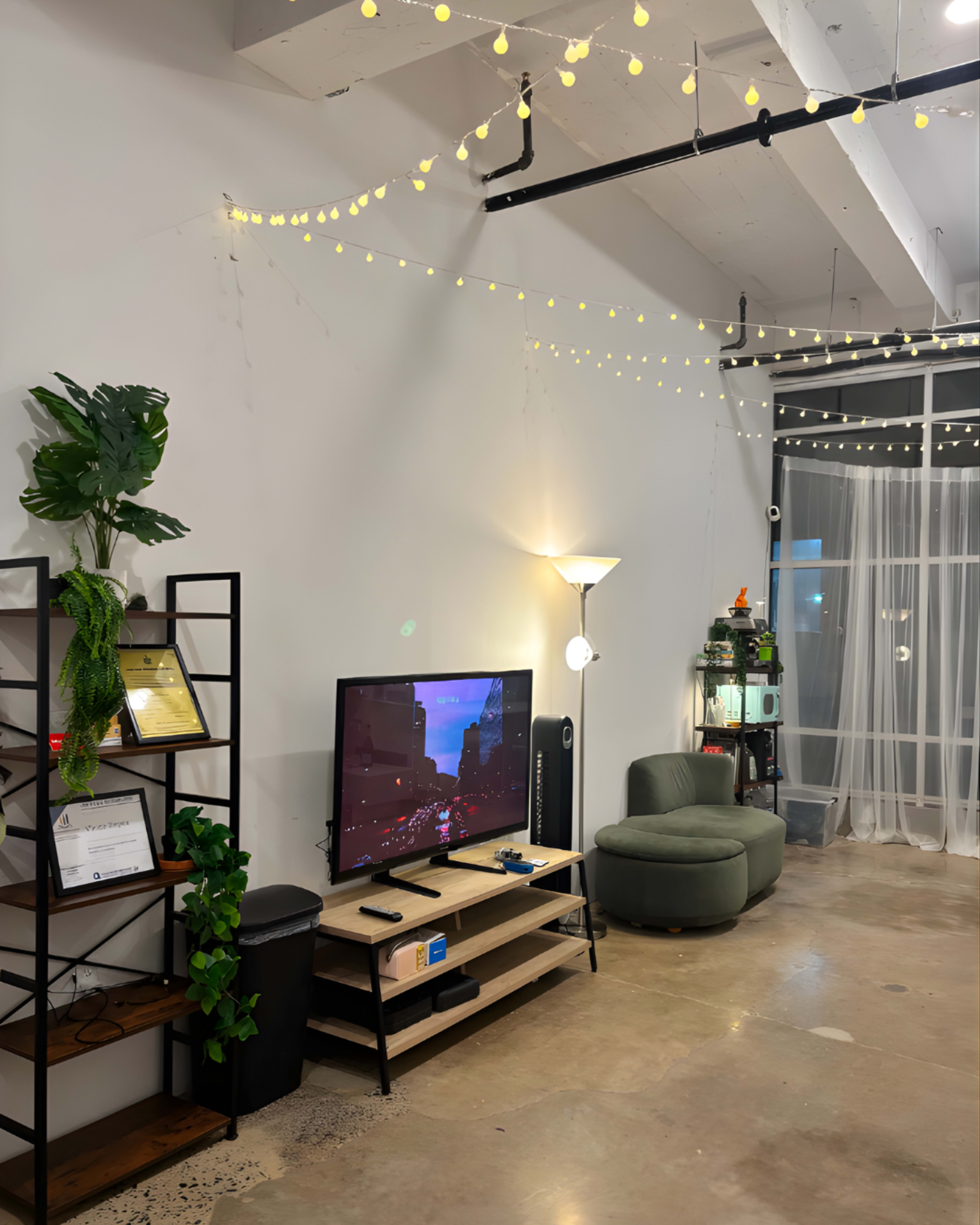 The image shows a modern living space with a TV mounted on the wall, a green sofa, a bookshelf, and string lights overhead.