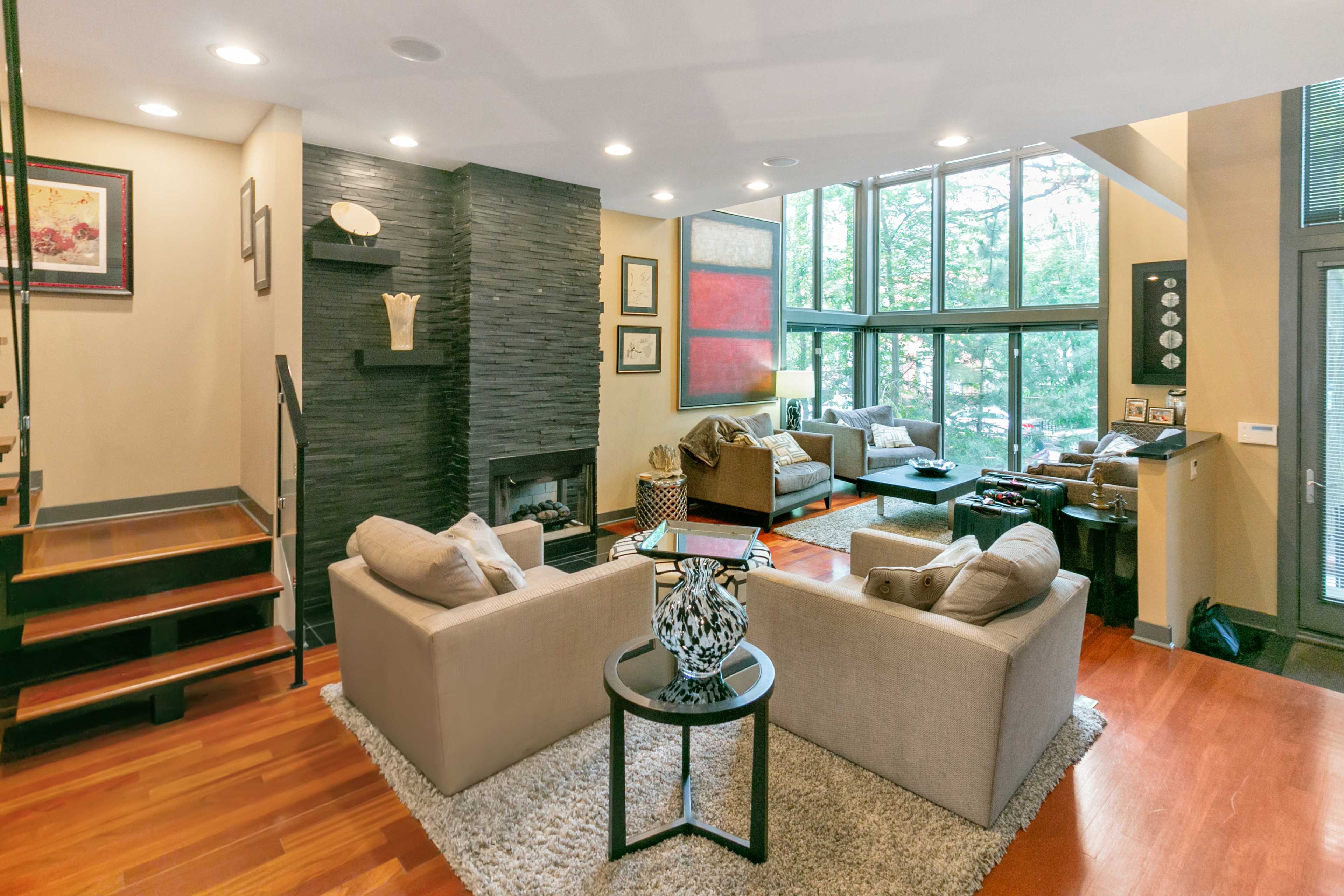 A modern living room features two sofas facing each other, a stone fireplace, large windows, and a staircase.