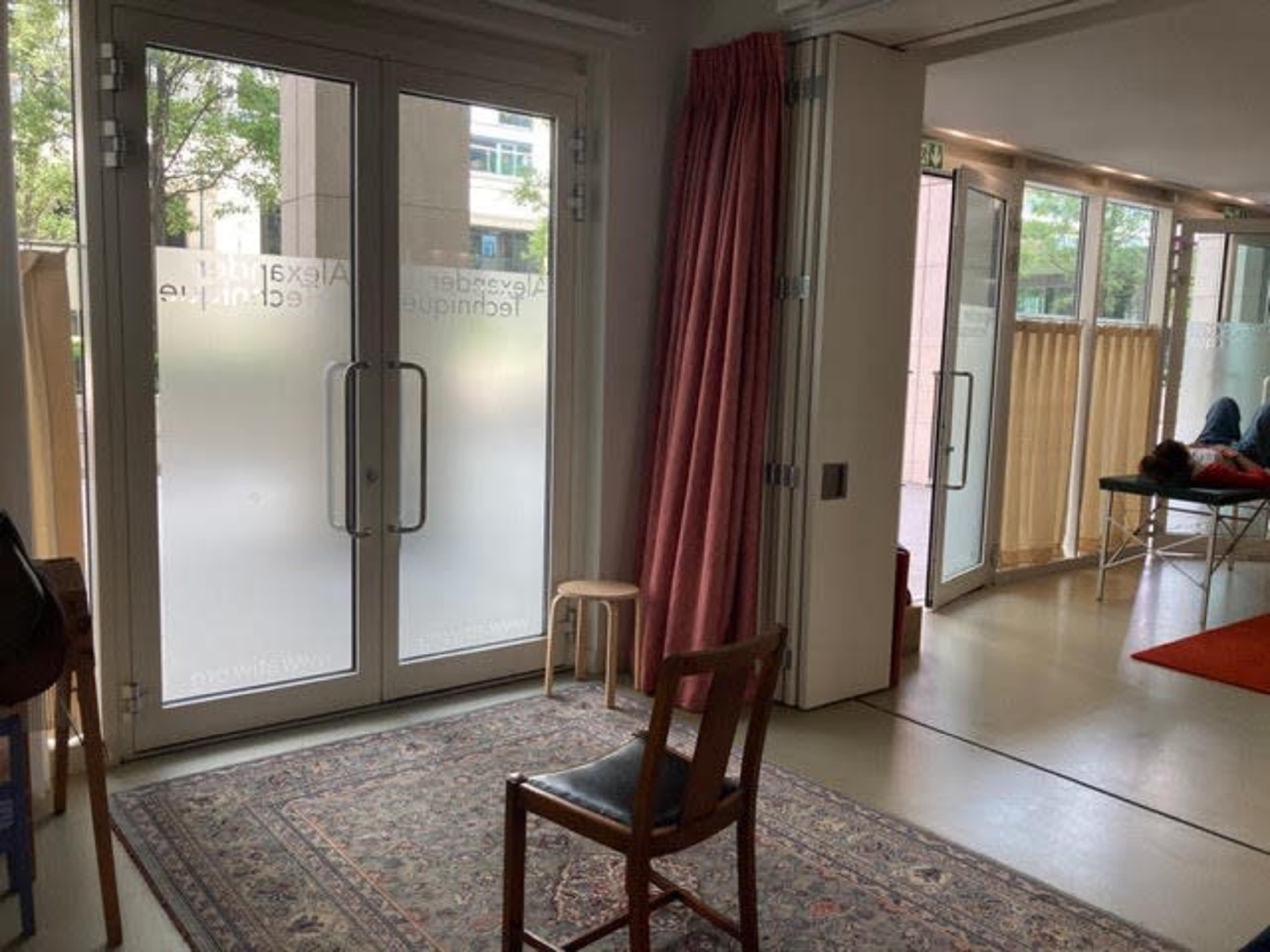 The interior of a room features a wooden chair on a rug, with large glass doors leading outside and a relaxation area with a person reclining on a table.