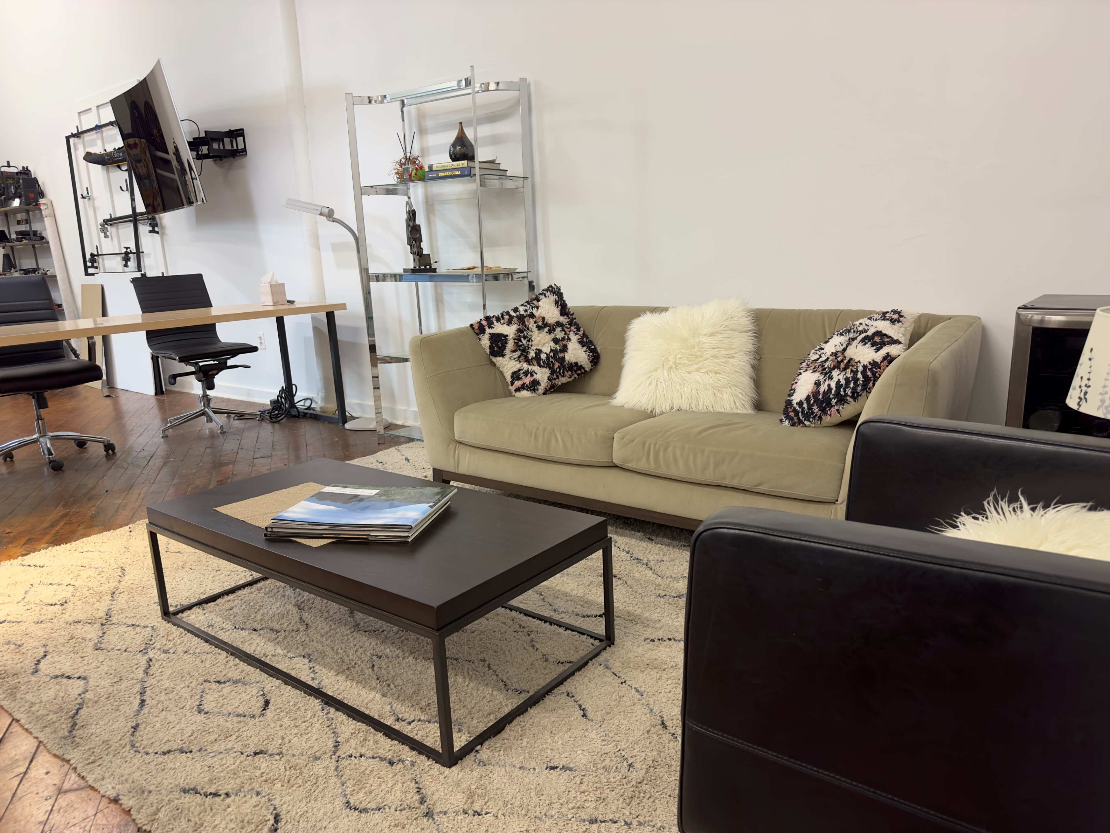 A modern seating area with a beige couch, two black chairs, a coffee table, and workspaces in the background.
