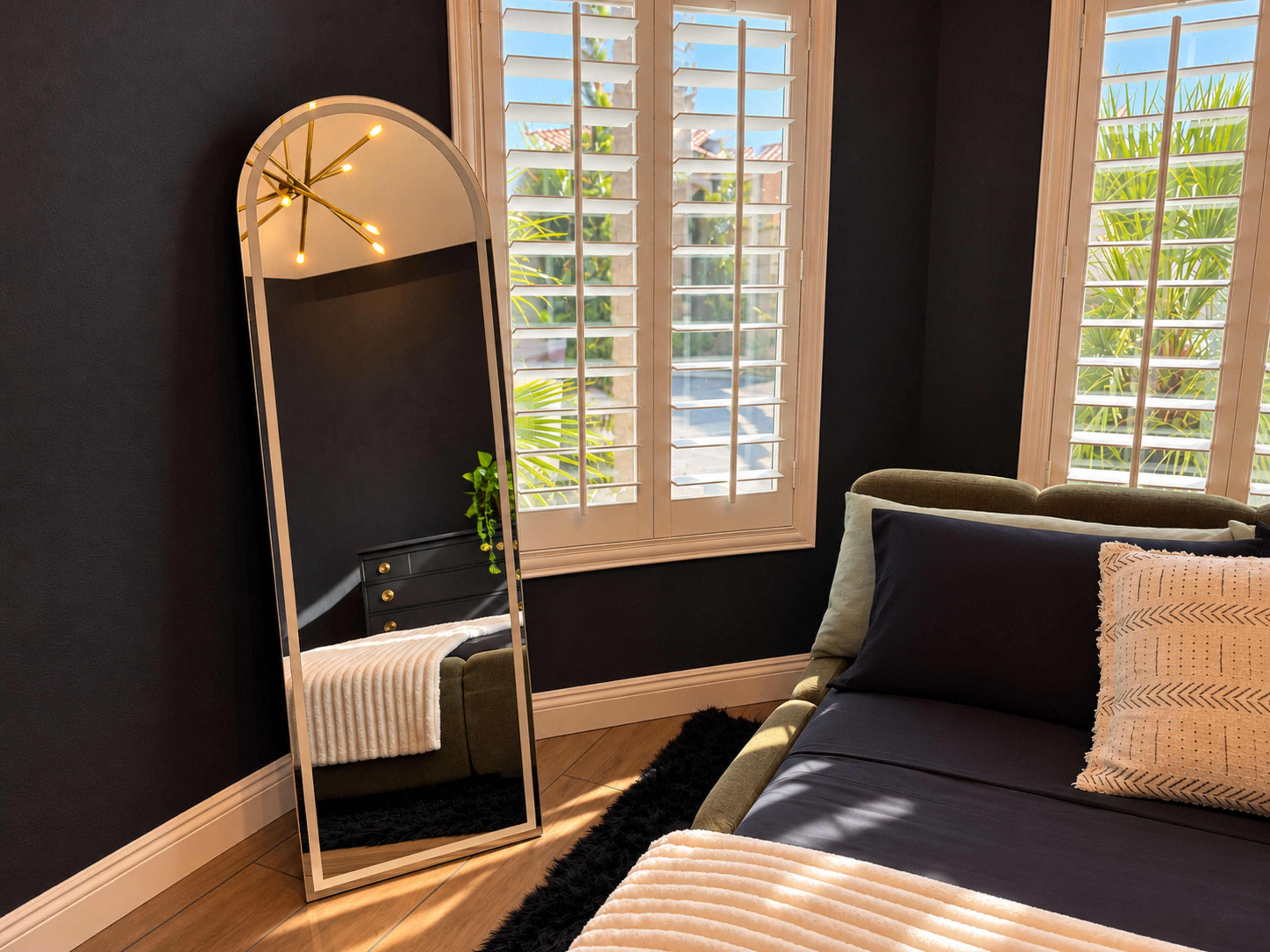 A large arched mirror reflects a well-lit corner of a stylish bedroom featuring a green sofa and window with plantation shutters.