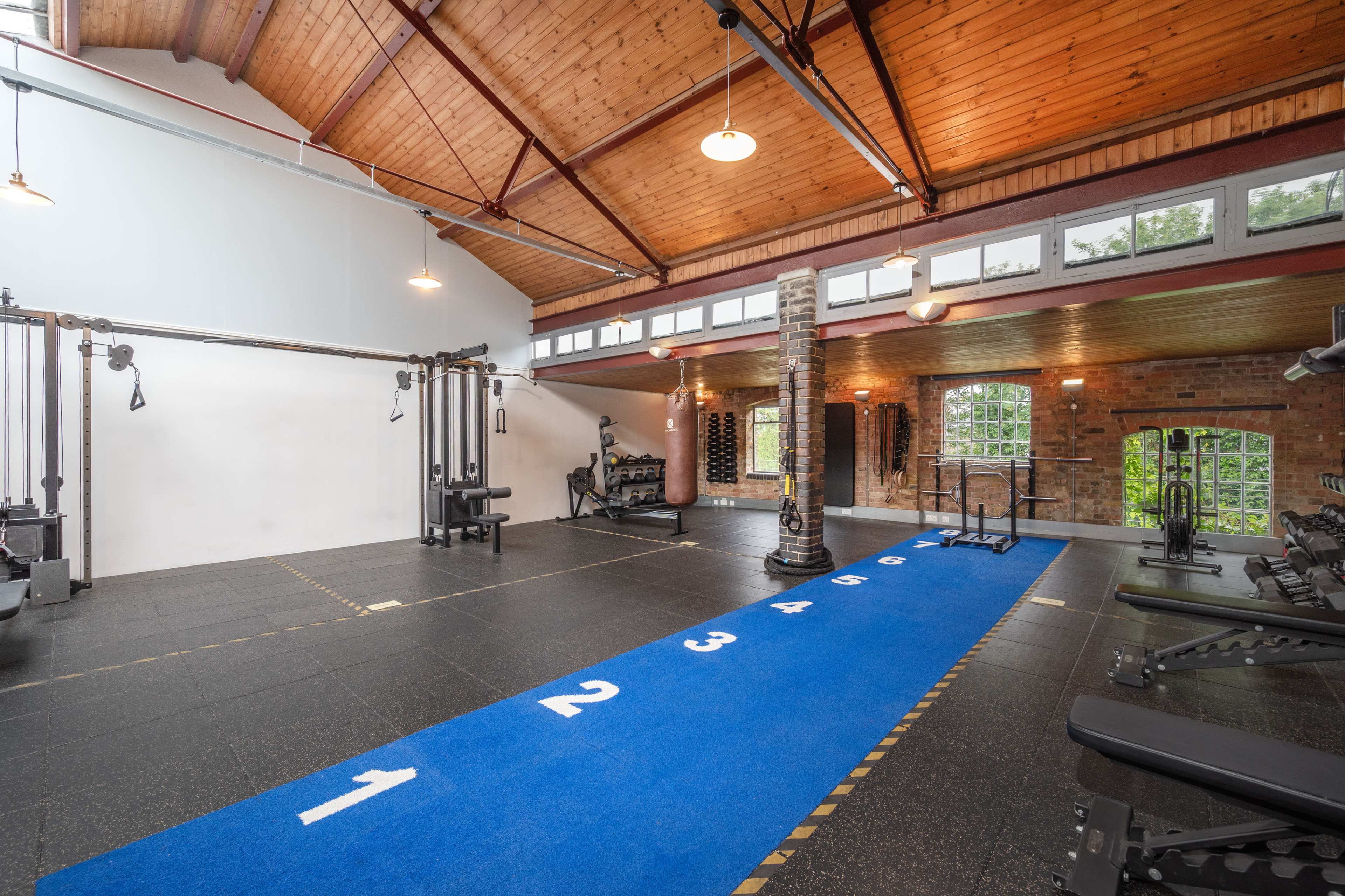 The image shows a spacious gym with a blue workout turf marked with numbers, surrounded by various exercise equipment and exposed brick walls.