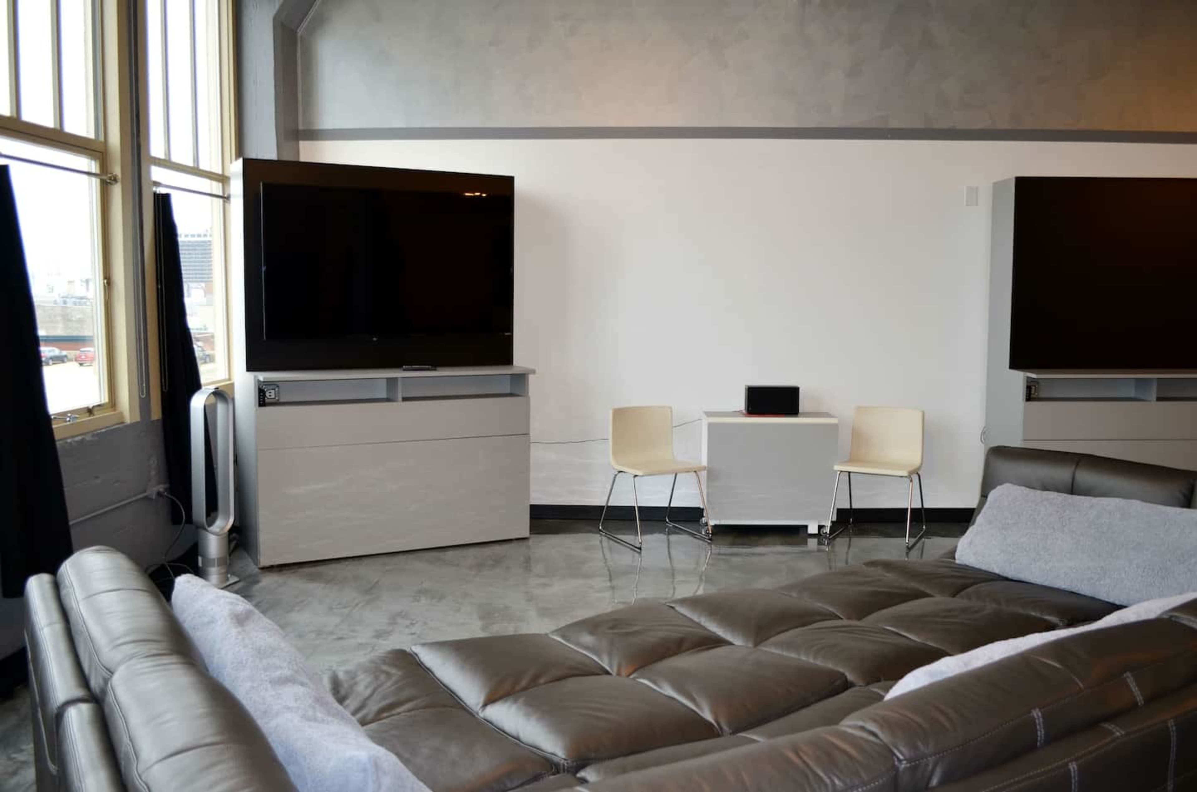 A modern living room features a gray sofa, two bare wooden chairs, a small table, and two large flat-screen TVs mounted on a light-colored wall.