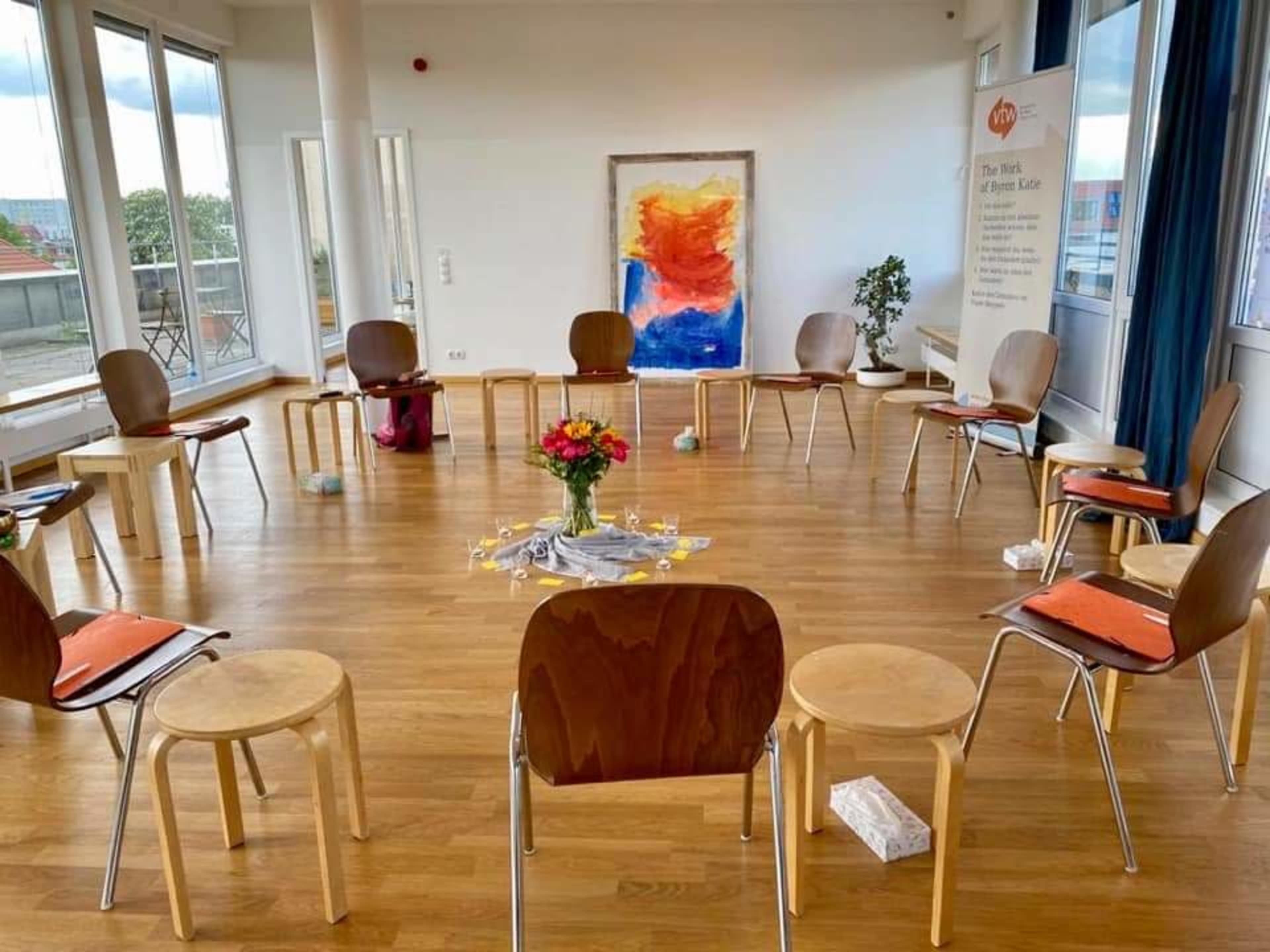 A circular arrangement of wooden chairs and a small table with flowers is set in a brightly lit room with large windows.