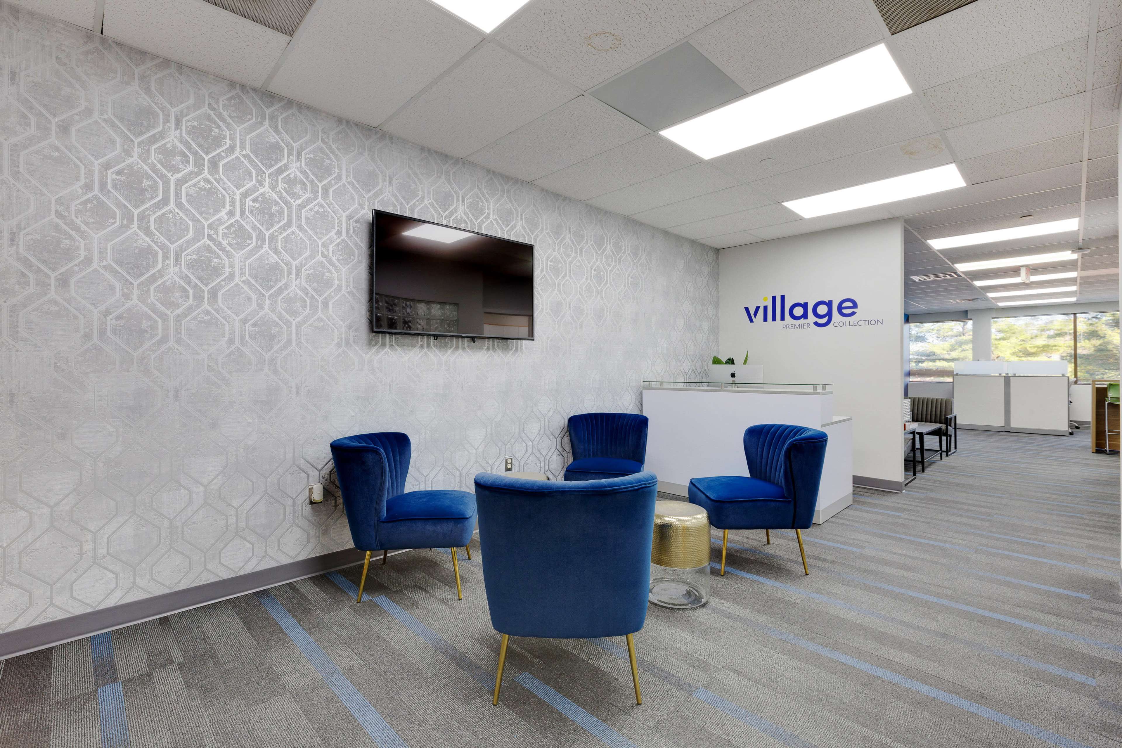 A modern office reception area with four blue chairs arranged around a small table, a wall-mounted TV, and the word "village" displayed on a white wall.