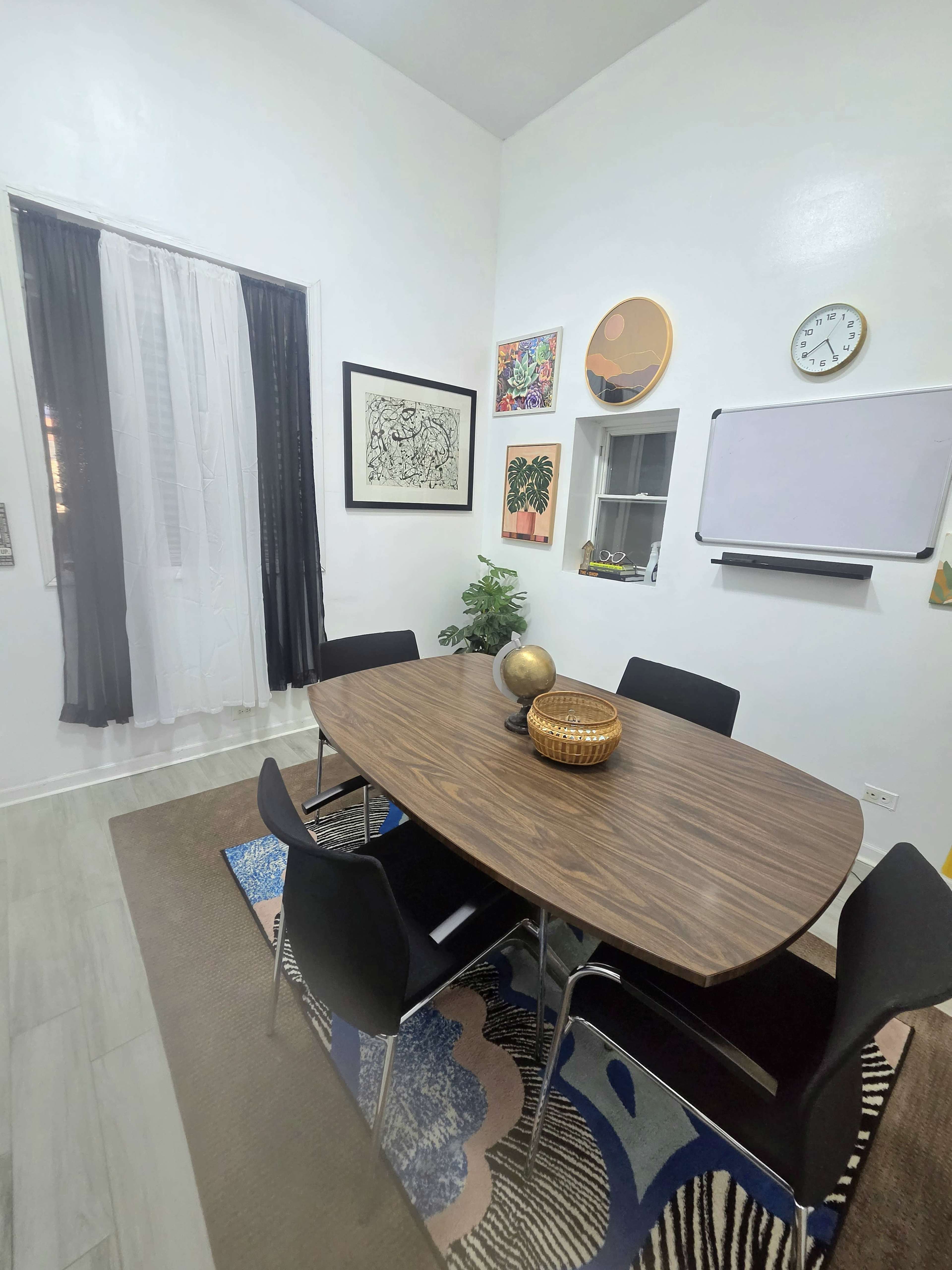 The image shows a small, well-lit room with a round wooden table surrounded by black chairs, artwork on the walls, and a large window with white curtains.