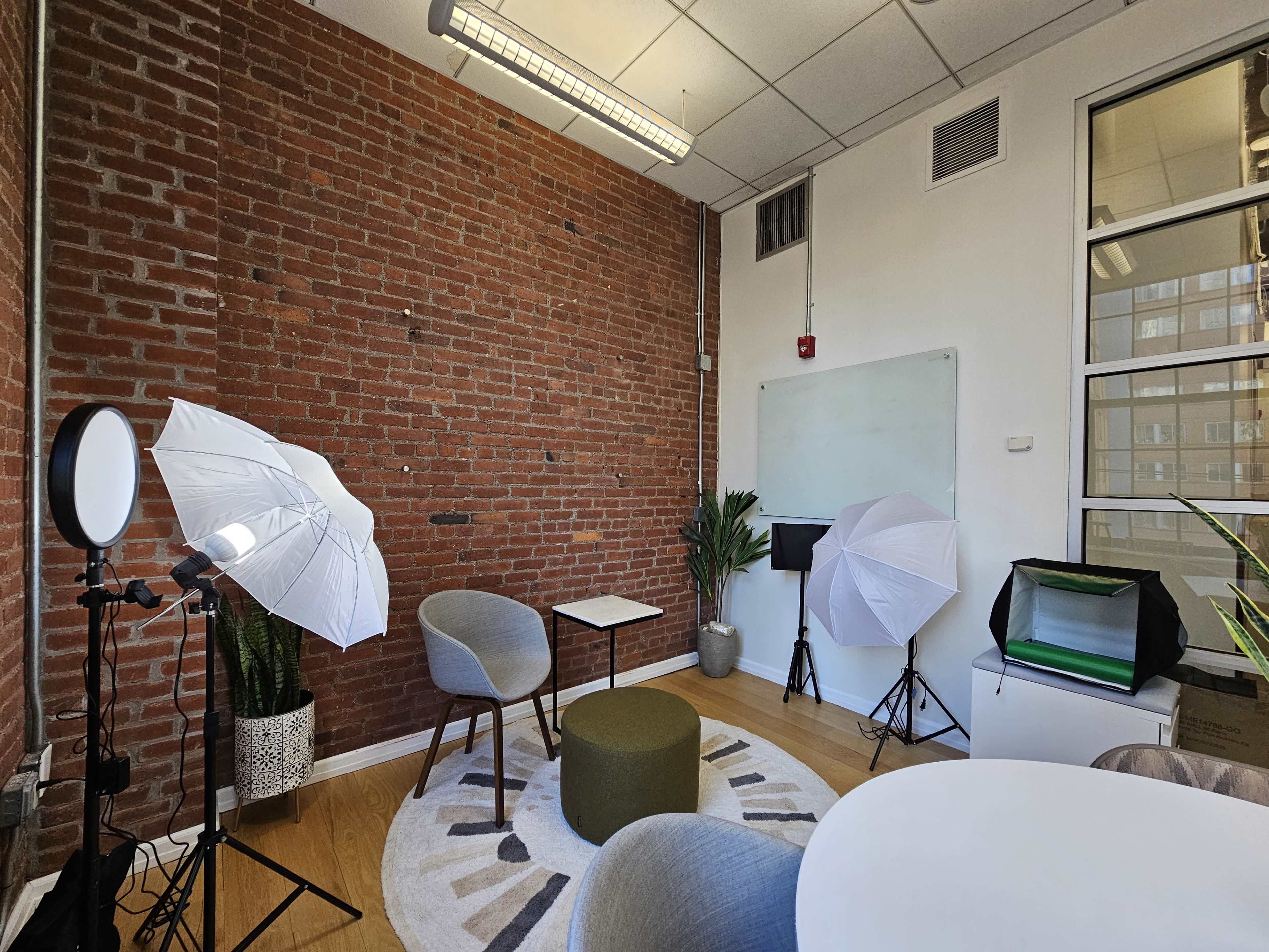 The image shows a small studio or office space with exposed brick walls, two chairs, a table, a round area rug, and professional lighting equipment set up for photography or video.