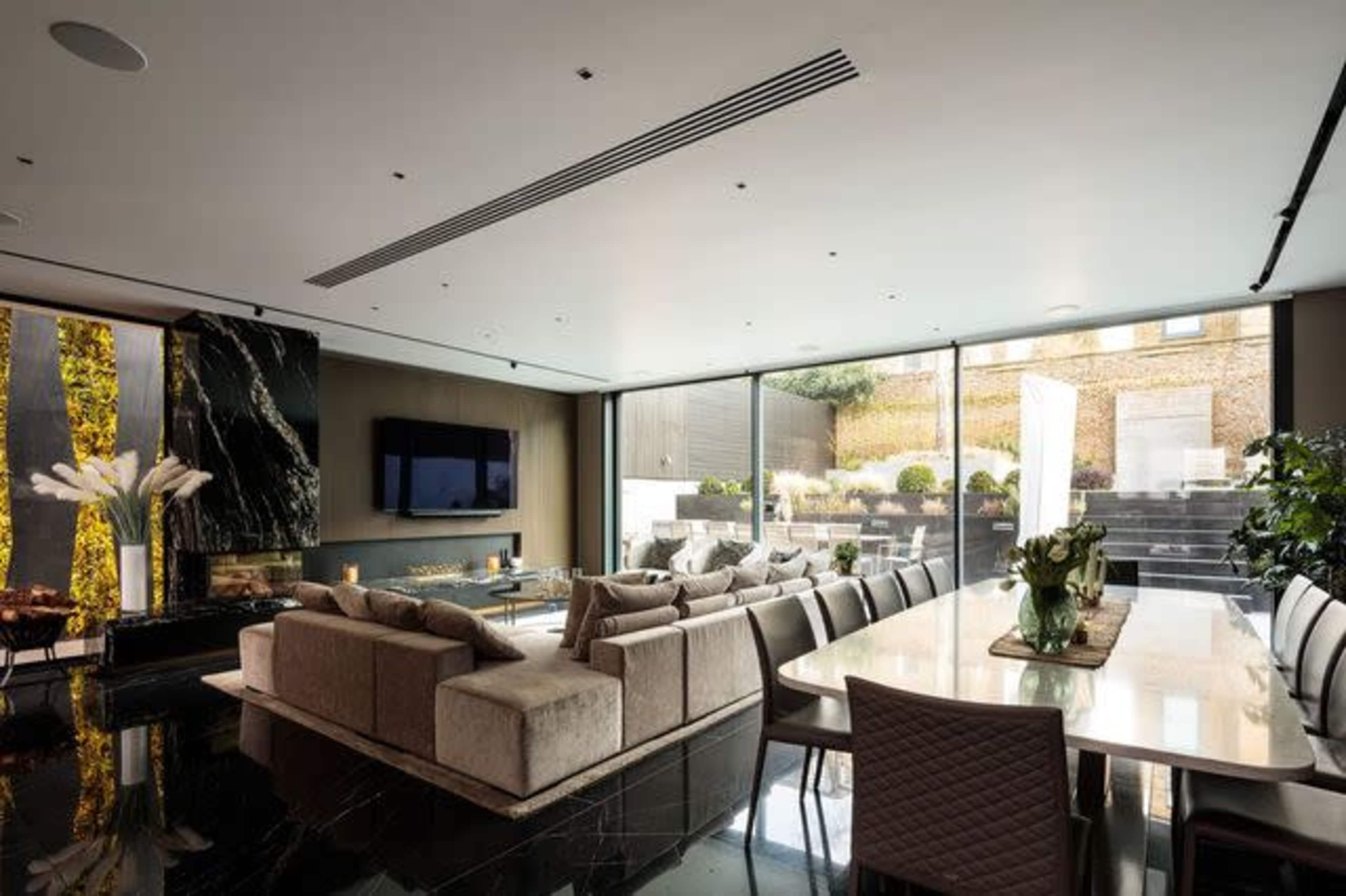 The image shows a modern living room with a large sectional sofa, a dining table, and floor-to-ceiling windows offering a view of an outdoor patio.