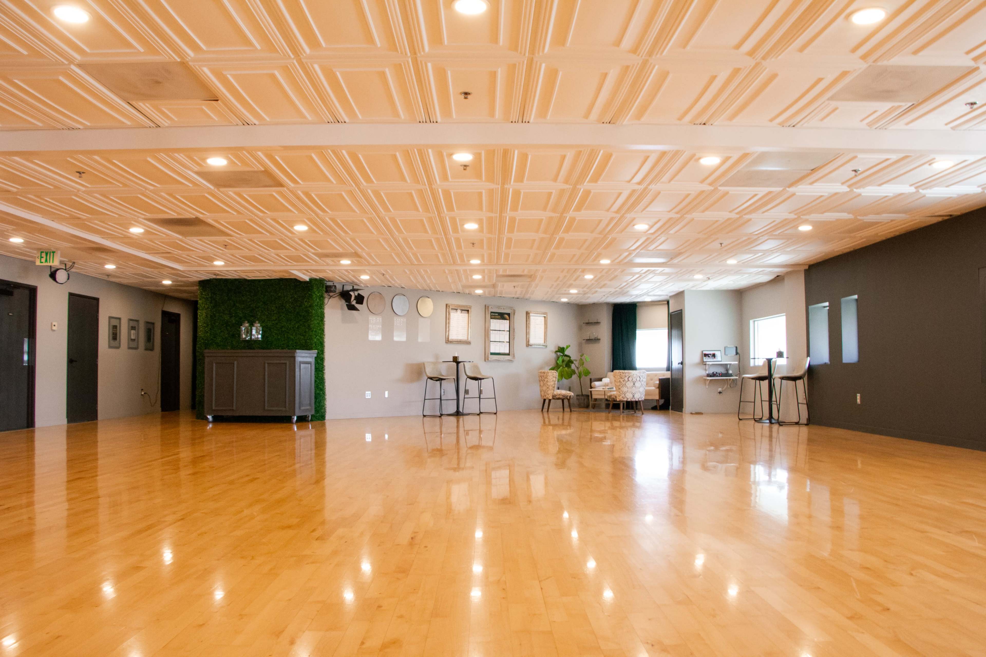 The image shows a spacious, well-lit studio with glossy wooden floors and a decorative ceiling, featuring a seating area and plants against the walls.